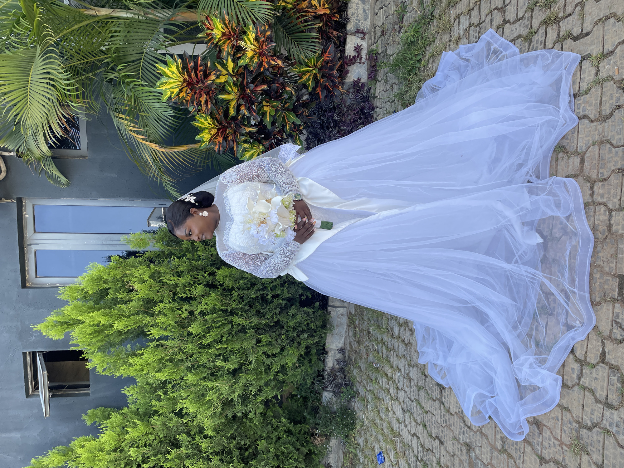 A woman in a white wedding dress stands on a paved surface. She is holding a bouquet of flowers. Behind her are green bushes and trees. A gray building with windows is to the left of the bushes. The location is possibly Jos, Nigeria.