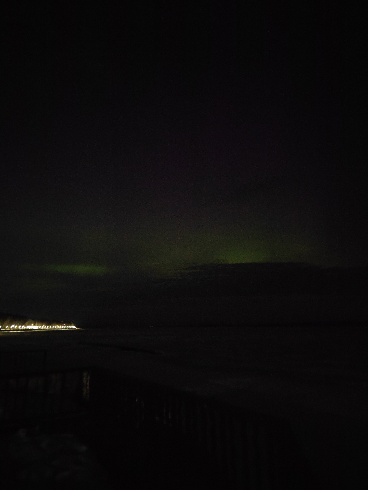 The image captures a night scene in Chaikovsky, Russia. The sky is predominantly dark with faint hints of green, possibly indicating the presence of the Northern Lights (Aurora Borealis). The lights of the city line the horizon, reflecting on the water. A dark fence or railing is visible in the foreground, adding depth to the composition. The overall environment suggests a quiet, cold night near a body of water.