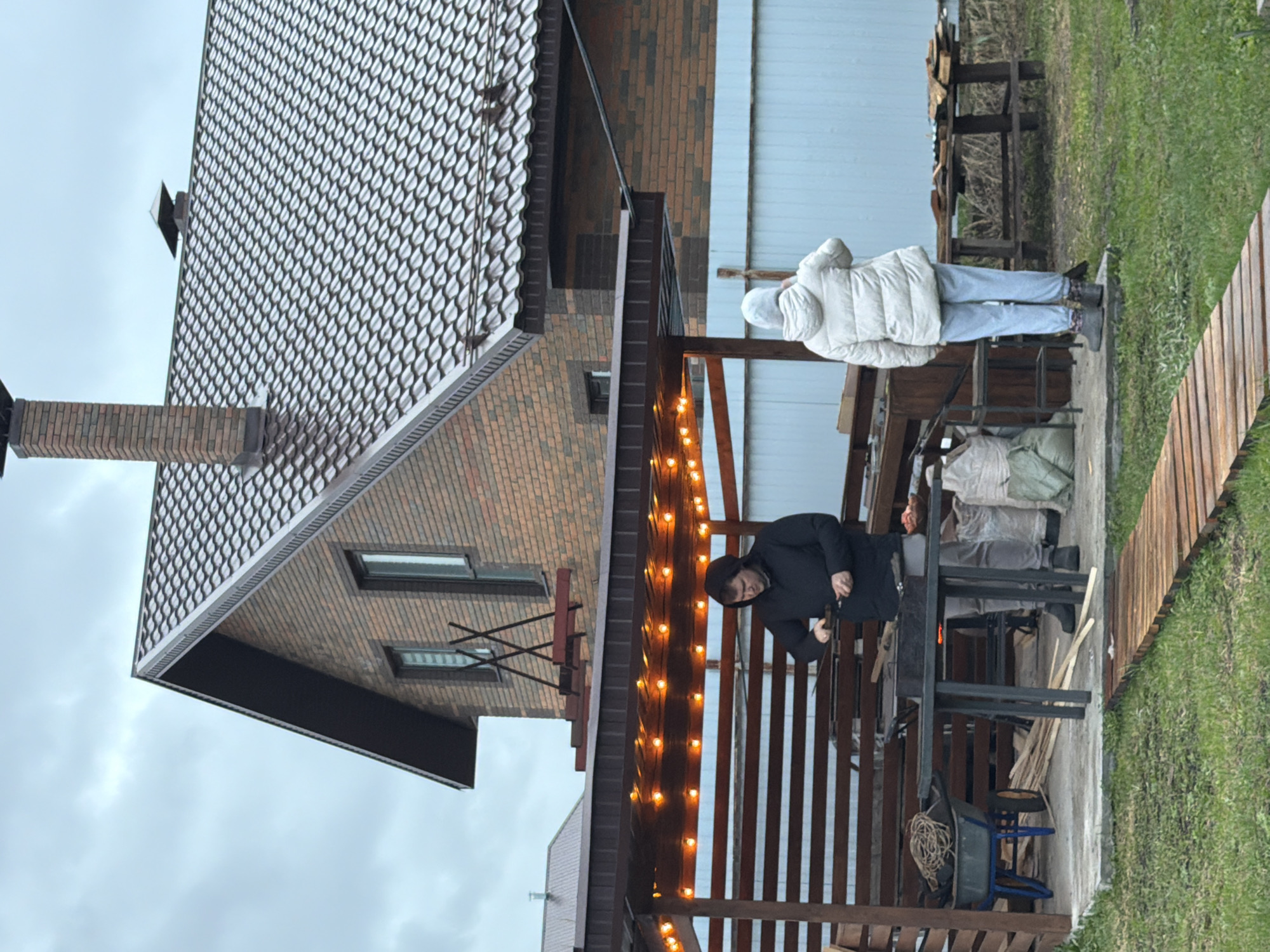 The image shows a scene in Большая Шильна, Russia. A two-story brick house with a patterned tile roof is visible on the left. To the right of the house is a wooden structure resembling a covered patio or deck, strung with warm-toned lights. Two people are present. One person, wearing a black coat and hat, is standing under the covered patio, appearing to be working on something. Another person, clad in a white puffer jacket and light-colored pants, stands outside of the patio. The surrounding environment includes grass and wooden planks, suggesting an outdoor setting.