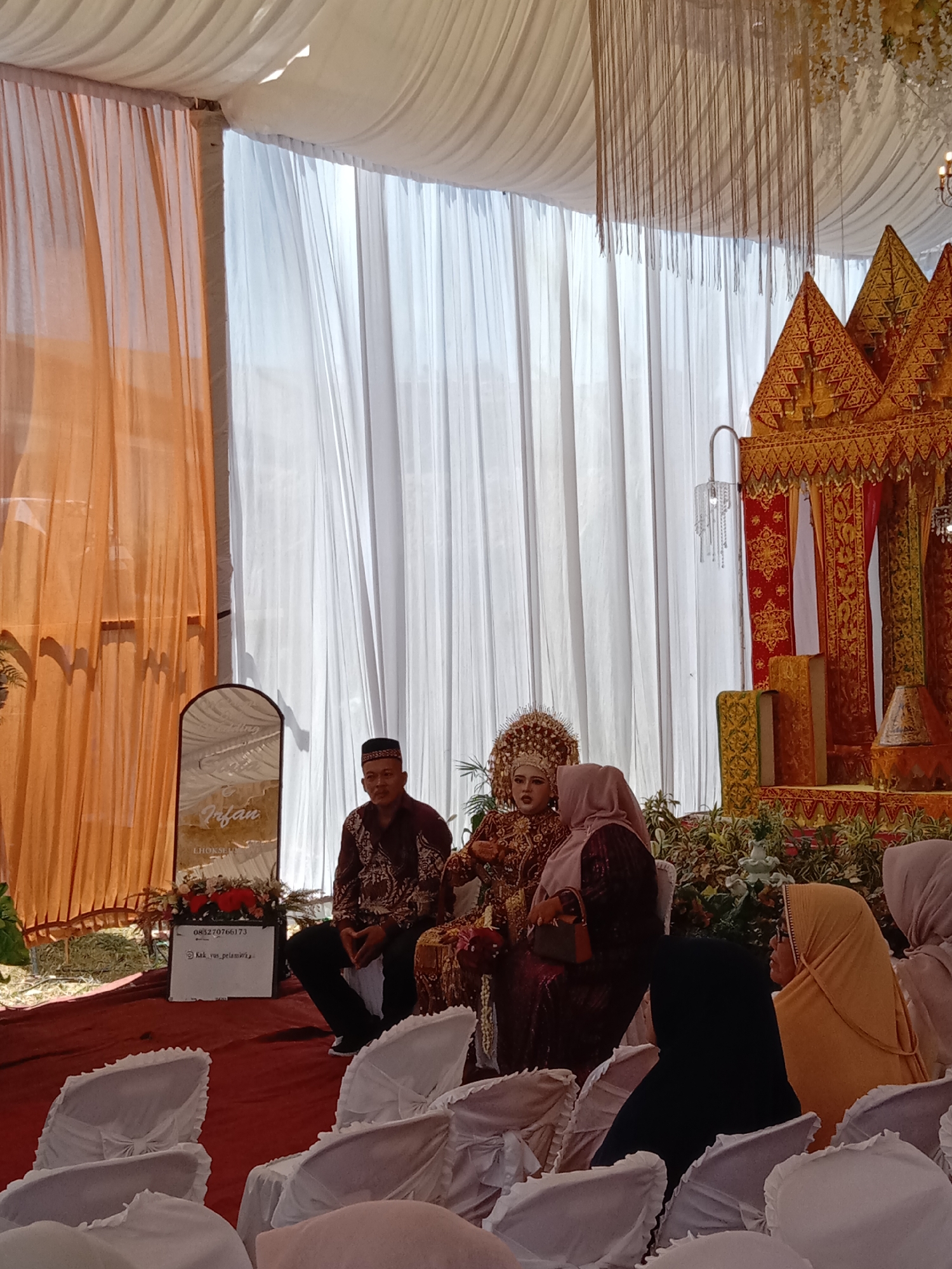 The image captures a wedding scene in Lhokseumawe, Indonesia. A couple, adorned in traditional attire, sits on a decorated platform. A man wearing a patterned shirt and cap sits to the left of the couple. Several women, dressed in hijab, are seated in the foreground, their backs to the camera. White chair covers adorn the seating area. Behind the couple is a decorated structure in gold and red tones. A sign with the name "Irfan LHOKSEUMAWE" is visible to the left. Drapes of orange and white fabric form a backdrop. The ground is covered with a red carpet.