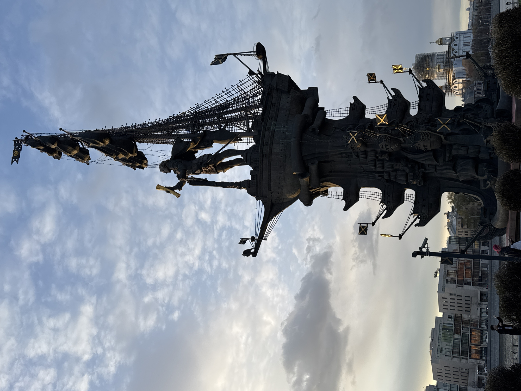The image depicts the Peter the Great Statue, a monument located on the Moskva River in Moscow, Russia. The statue, rendered in dark metal, features a large ship with sails and rigging. Peter the Great stands on the ship's deck, holding a scroll and looking towards the horizon. The background shows a cityscape with buildings and cloudy sky.