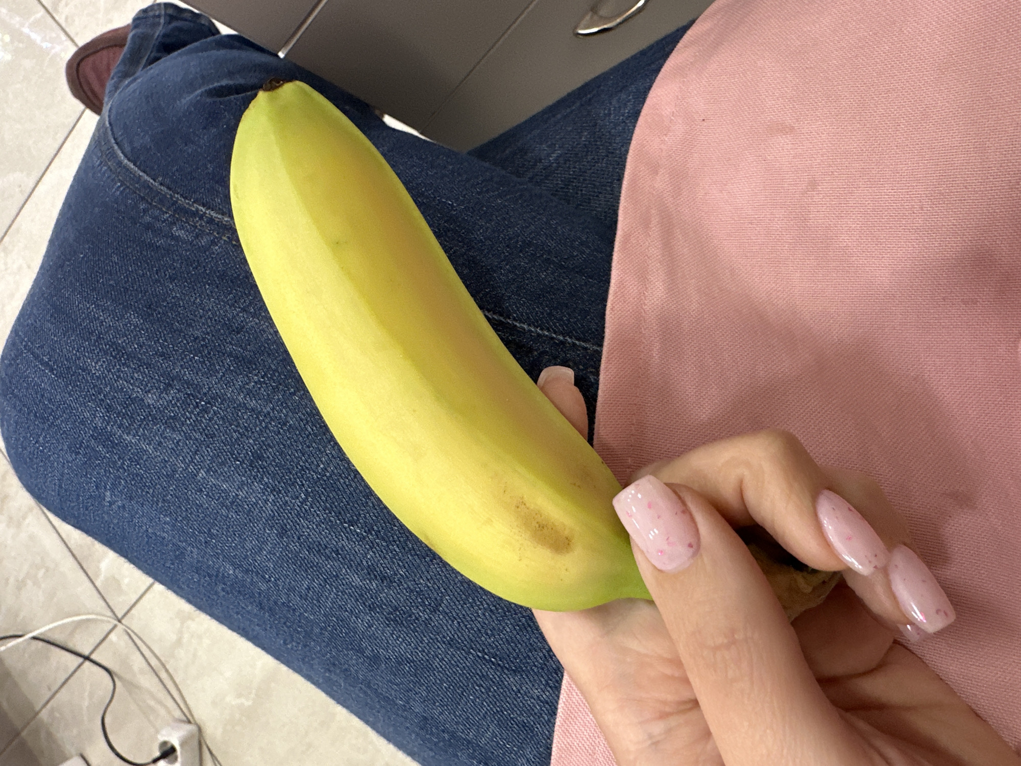 A person is depicted in a high-angle, close-up shot, seated indoors. The main subject is a left hand holding a single ripe yellow banana. The hand is manicured with oval-shaped nails painted in a light pink polish adorned with small, darker pink specks. The banana shows a small brown spot on its skin and has a greenish tip.
The person is wearing dark blue denim jeans, visible on their legs, and a soft, textured pink garment, likely a top or apron, covering part of their lap. A dark brown shoe is partially visible on the tiled floor near the top left. The floor is covered with light-colored tiles. In the bottom left corner, electrical wires and a white plug are visible on the floor, suggesting a power outlet nearby. A metallic edge, possibly belonging to a laptop or a piece of furniture, can be seen in the upper middle section.
The scene conveys a casual, everyday moment in an indoor setting, potentially a home, office, or public space. No specific time of day or weather conditions are discernible. No emotions are explicitly conveyed, and no visible text appears in the image. The location context is Sofia, Bulgaria, though no specific visual cues within the image confirm this.