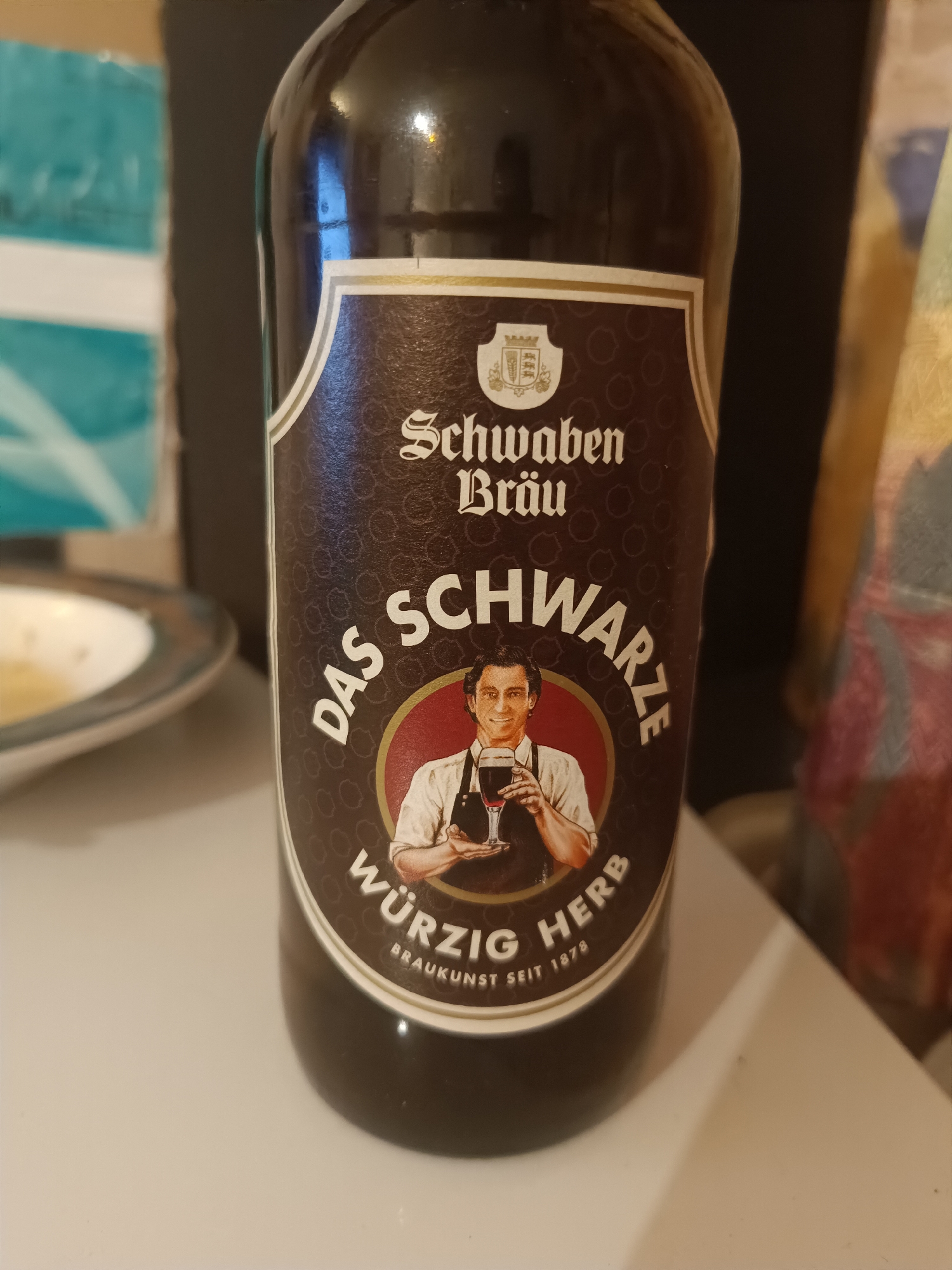 A close-up shot of a dark glass bottle, labeled "Schwaben Bräu DAS SCHWARZE WÜRZIG HERB BRAUKUNST SEIT 1878." The label features a circular image of a man holding a dark beer. Schwaben Bräu is a brewery founded in 1878 in Seewald, Germany.