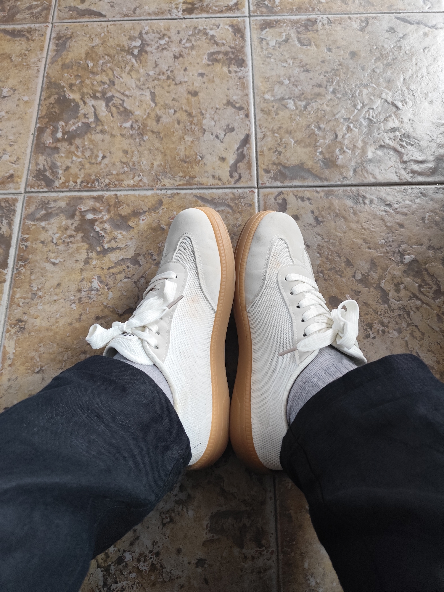 This is a high-angle, full-shot photograph of a person's feet wearing white sneakers with gum soles and dark pants. The sneakers are a low-top style with white laces and mesh detailing on the sides. Gray socks are visible above the shoes. The person is standing on a tiled floor with a mottled brown and beige pattern, with visible grout lines. The image appears to be taken indoors, possibly in a home or a commercial space, but there are no elements in the image that would specifically place it in Mtskheta, Georgia.
