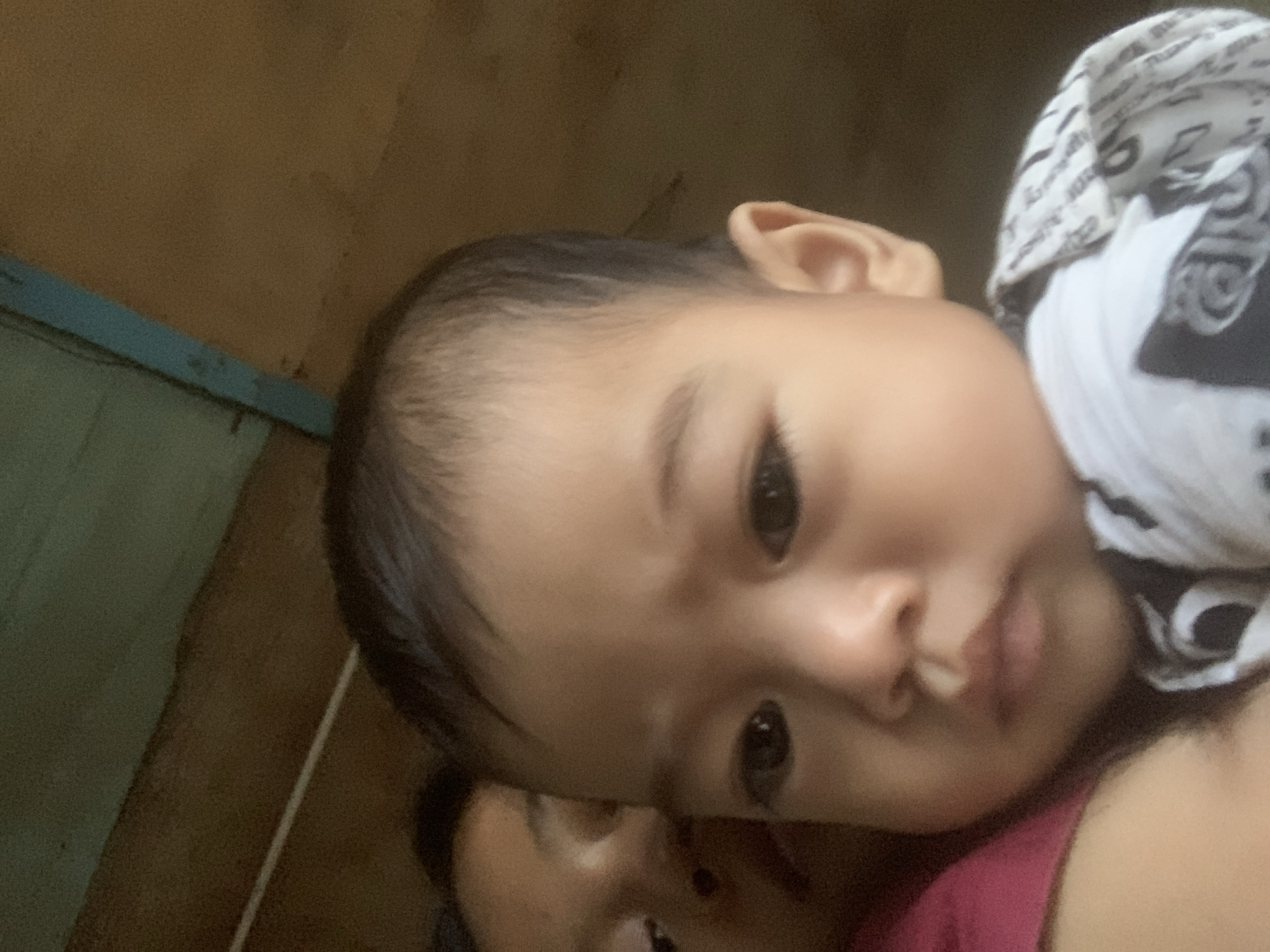A baby with dark hair lies against a person wearing a magenta shirt. The baby is wearing a white and black patterned cloth draped over their shoulder. The background shows a plain wall. The location is Sungai Meriam, Indonesia.