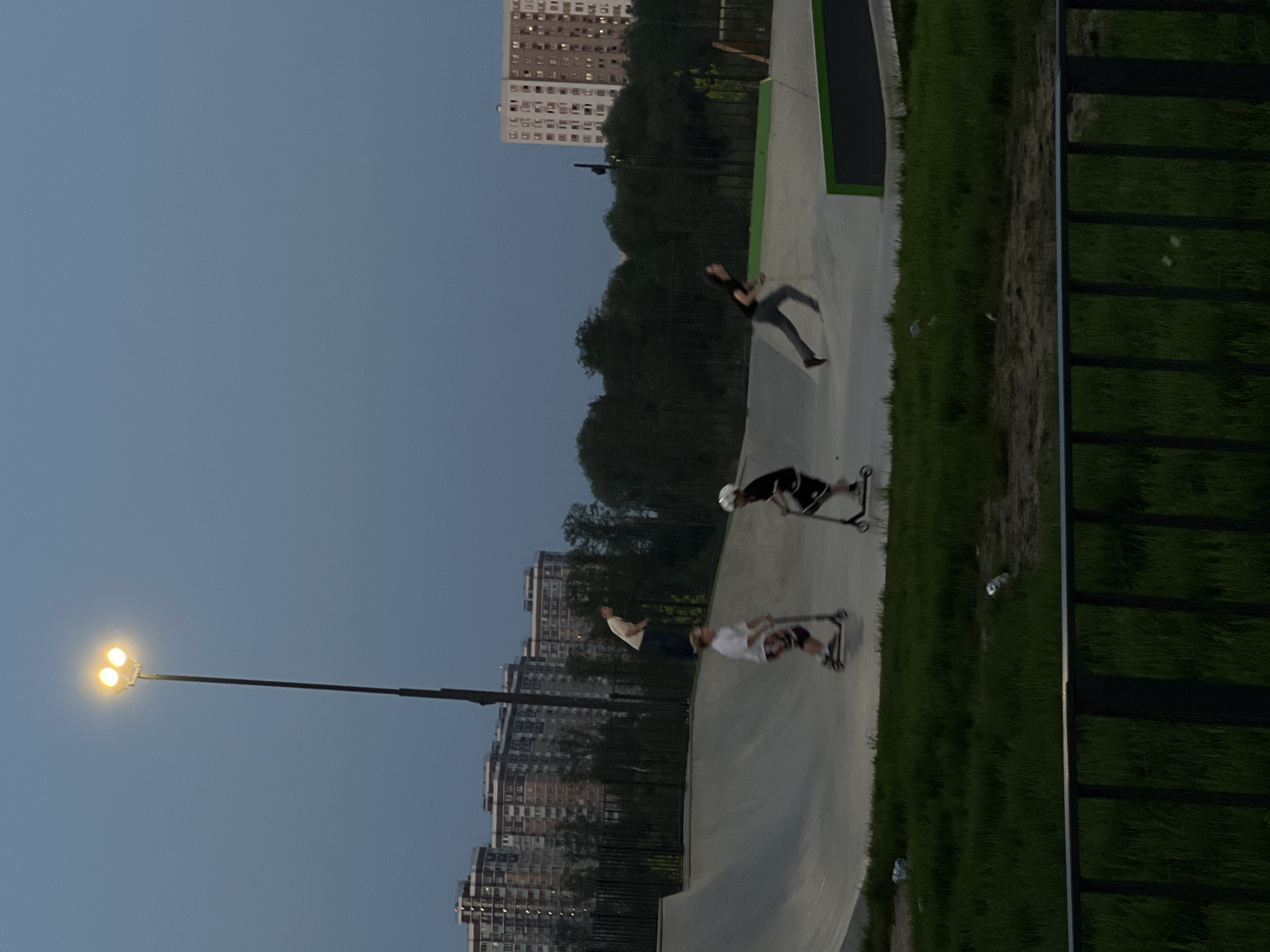 The image depicts a skate park in Moscow, Russia, at dusk. A streetlamp illuminates the scene. Four individuals are visible; three are riding scooters on the concrete ramps of the park, while the fourth is running along the edge of a ramp. Apartment buildings are visible in the background. Adjacent to the concrete of the skate park is a strip of grass, alongside a green fence.