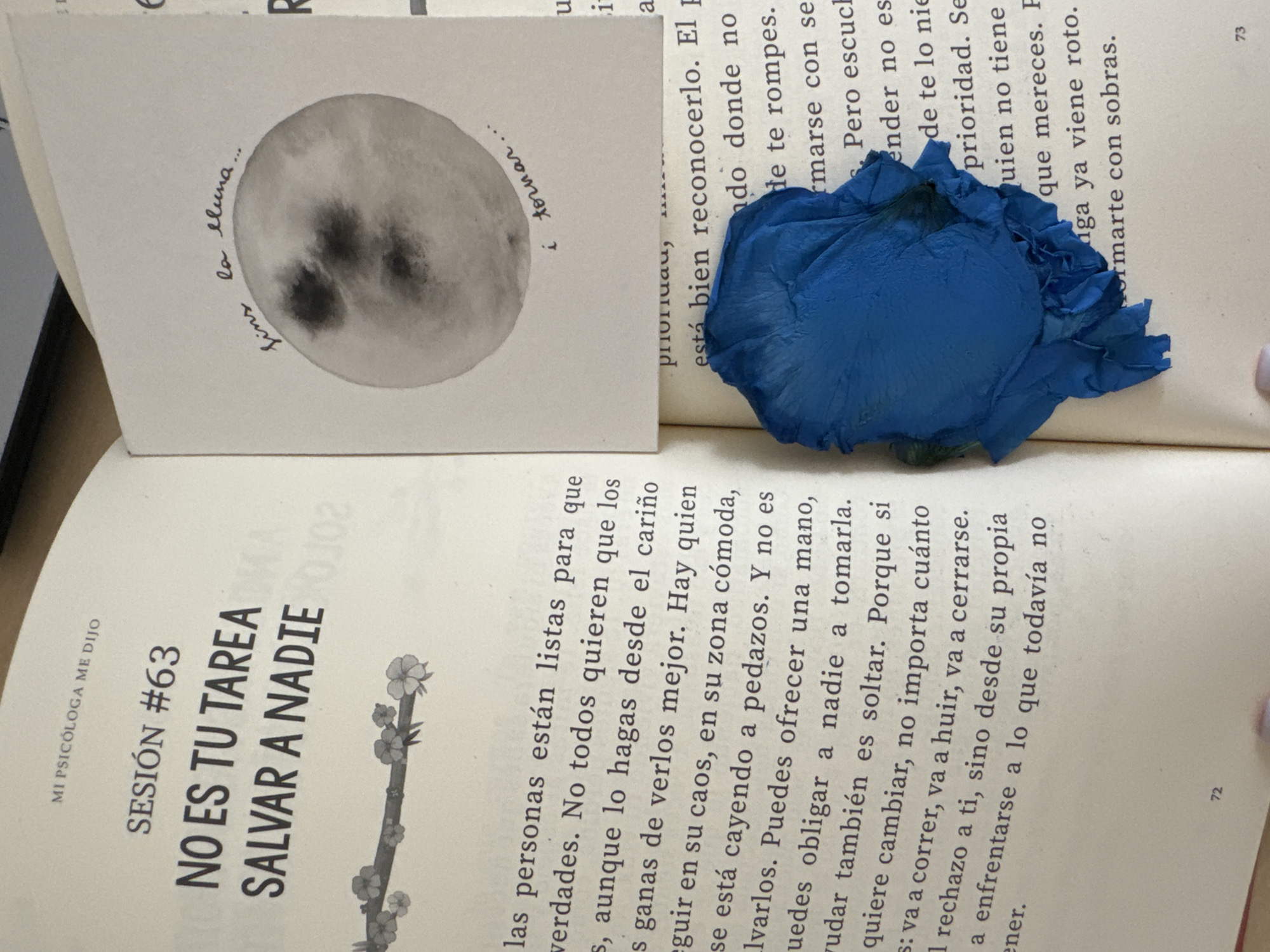 The image shows an open book with text and a small drawing. The book is on a wooden surface, likely a table. On the left page, the text reads "MI PSICÓLOGA ME DIJO SESIÓN #63 NO ES TU TAREA SALVAR A NADIE". Below this, there is an illustration of a branch with flowers. The right page has handwritten text above a watercolor painting of the moon that says "fins la lluna..." and "i tornar...". A blue, crumpled object, possibly a dried flower petal or piece of fabric, is placed on the right page, partially obscuring the text which says "prioridad," and "está bien reconocerlo. El pando donde no de te rompes." Further text on the right page is partially visible and includes words like "rmarse con se." and "Pero escue." and "ender no es de te lo nie." and "prioridad. Se uien no tiene que mereces. P." and "ga ya viene roto. rmante con sobras." The page number "72" is visible at the bottom of the left page, and "73" is visible at the bottom right. There is no information in the image to suggest a location in Murcia, Spain.