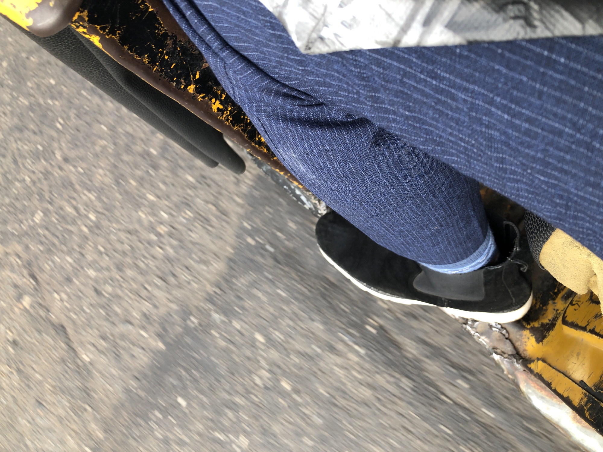 The image shows a close-up view of a person's legs and feet from a seated position, likely in a vehicle moving through the city of Lagos, Nigeria. The perspective is from inside a vehicle, looking down at the passenger's lower body and the vehicle's interior.
The person is wearing dark blue pinstripe trousers and black suede slip-on shoes with white soles. One leg is slightly bent, with the foot resting on what appears to be a textured, possibly metal, surface. A black, padded armrest or seat back is visible behind the person's legs. Part of the vehicle's yellow and black hazard warning striping is also visible on the lower right side of the frame, suggesting the vehicle might be a bus, van, or perhaps a commercial tricycle (Keke Napep) common in Lagos.
The background is blurred due to motion, showing a grey, textured surface that is consistent with a paved road in Lagos. There are small white and light-colored specks scattered across the road surface, which could be debris or variations in the asphalt. The overall impression is of movement and a typical urban streetscape in Nigeria. The angle of the shot and the motion blur suggest the image was taken while the vehicle was in transit.
