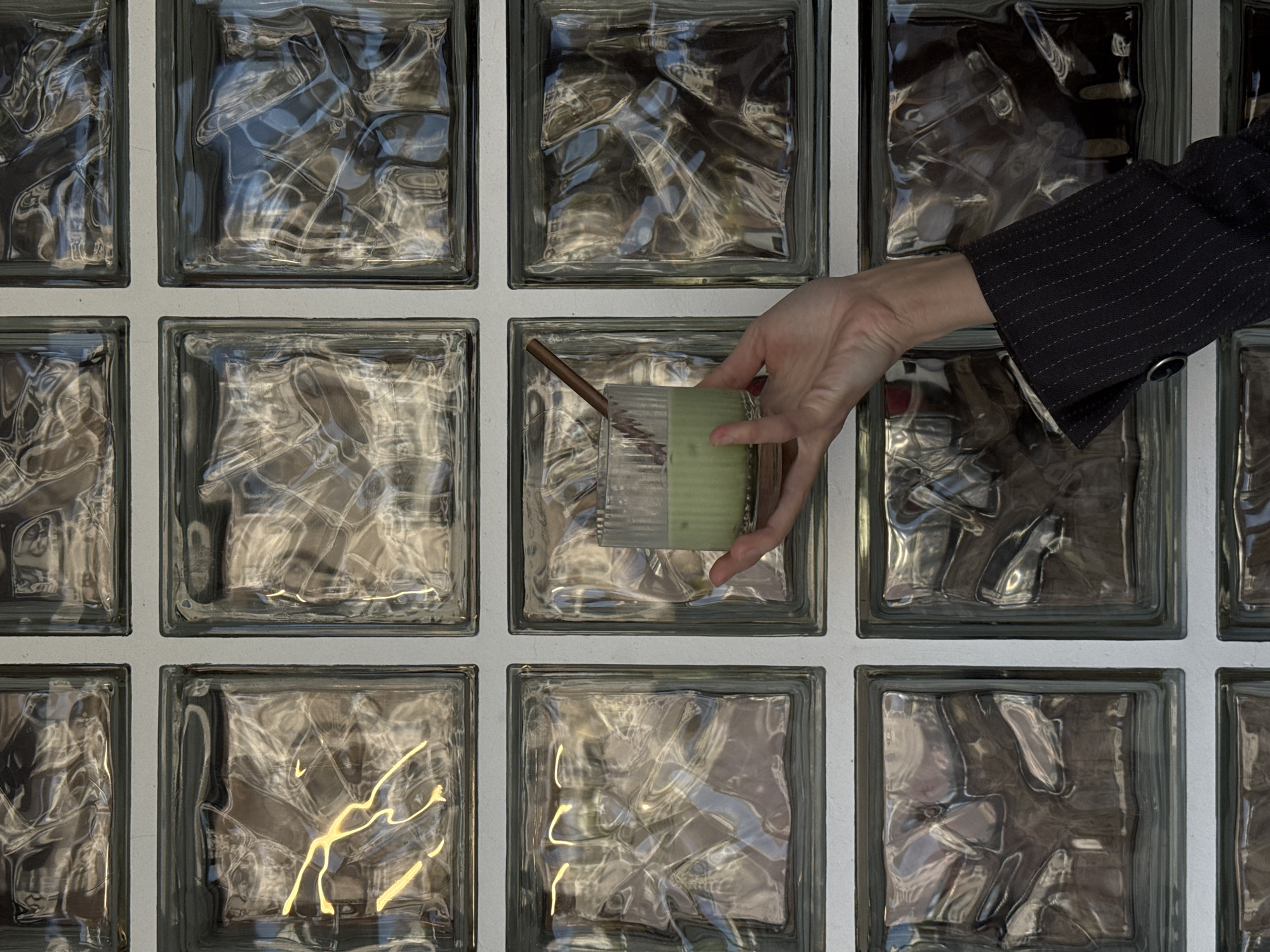 A close-up, slightly high-angle shot shows a person's hand holding a light green beverage in a clear glass with a straw in front of a glass brick wall. The person wears a dark pinstripe suit jacket. The glass brick wall, possibly a structural design element, consists of twelve visible square bricks. Each brick has a rippled, textured interior. The location is Bar, Montenegro, though it is not possible to determine the exact location within Bar from this image alone. The action is someone presenting or holding the beverage.