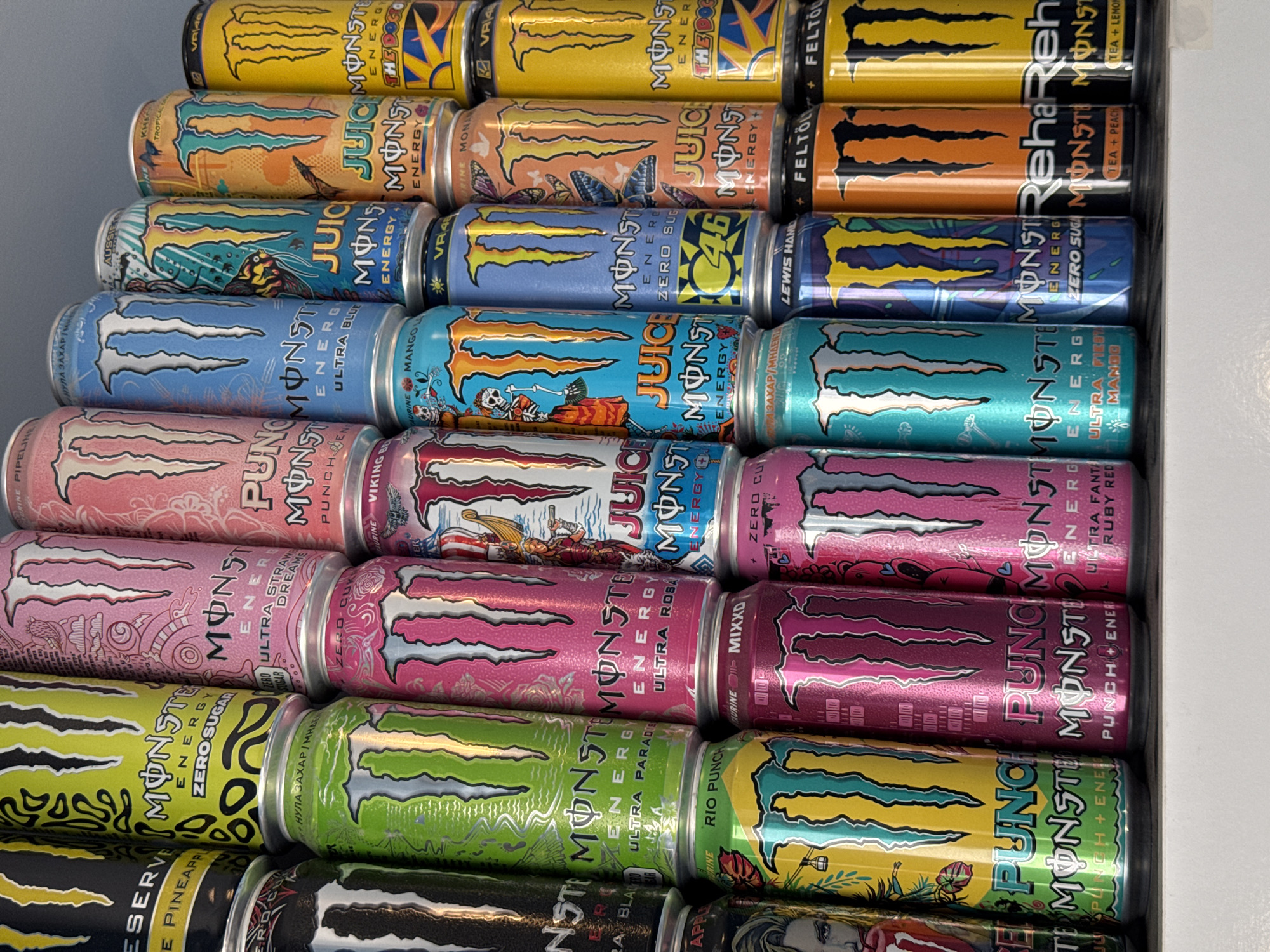The image shows a close-up view of a refrigerated display filled with various flavors of Monster Energy drinks. The cans are stacked tightly, with several rows visible. The flavors represented include original Monster Energy, Monster Rehab Tea + Lemon, Monster Rehab Tea + Peach, Monster Ultra Rosa, Monster Ultra Paradise, Monster Ultra Strawberry Dreams, Monster Ultra Fiesta Mango, Monster Lewis Hamilton Zero Sugar, Monster The Doctor, Monster Pipeline Punch, Monster Mango Loco, and Monster Aussie Style Lemonade. The lighting is bright and somewhat cool, typical of a refrigerated display. The cans feature a mix of colors and designs. The image doesn't provide sufficient information to identify the location as Burgas, Bulgaria.
