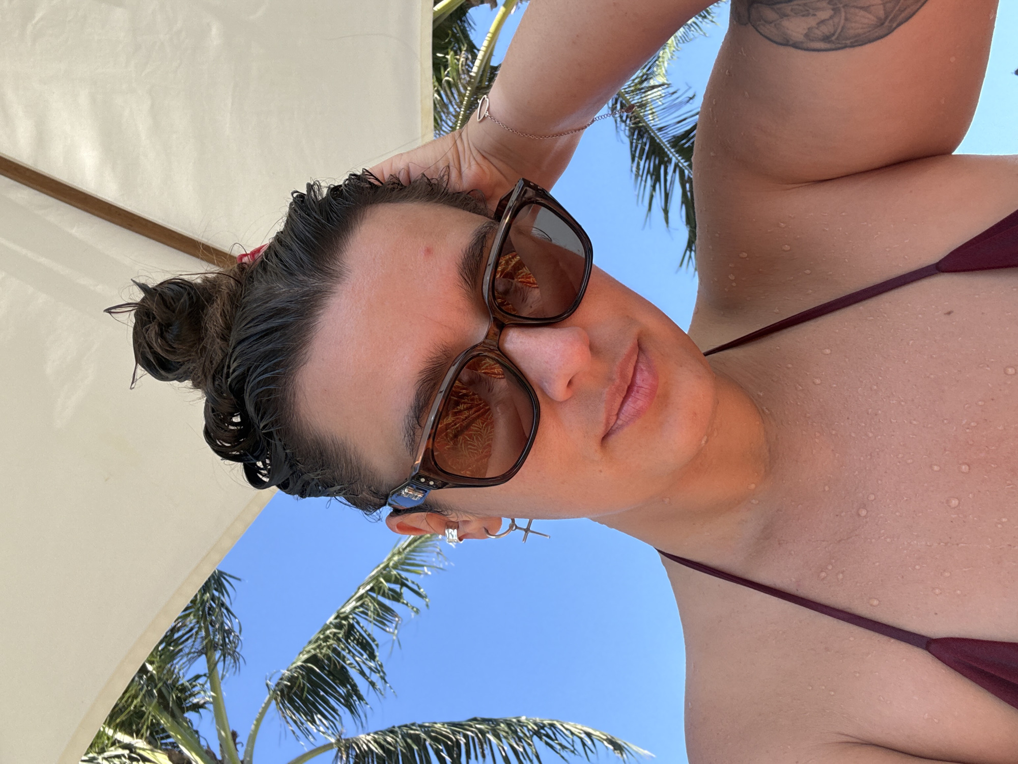 A woman in a burgundy bikini poses under a white umbrella on a sunny day. Palm trees are visible in the background against a blue sky. She wears brown sunglasses, silver earrings, and a silver bracelet. The woman's hair is wet and pulled back in a bun. The water droplets on her skin indicate she may have been swimming. The location is likely Keramas, Indonesia, implied by the presence of palm trees and tropical environment.