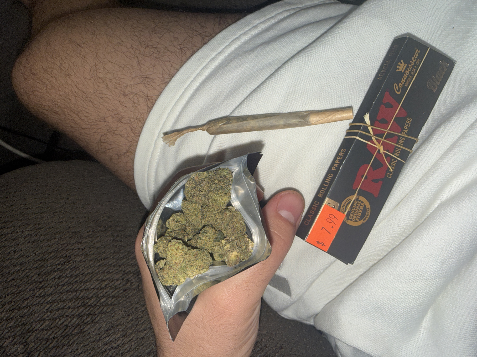 The image shows a person's lap, likely indoors. The person is wearing white clothing. On the clothing is a bag of what appears to be marijuana, a rolled joint, and a package of RAW Connoisseur Black King Size Slim rolling papers with tips, priced at $7.99. No indication of a location in Ocean City, United States of America, is visible.