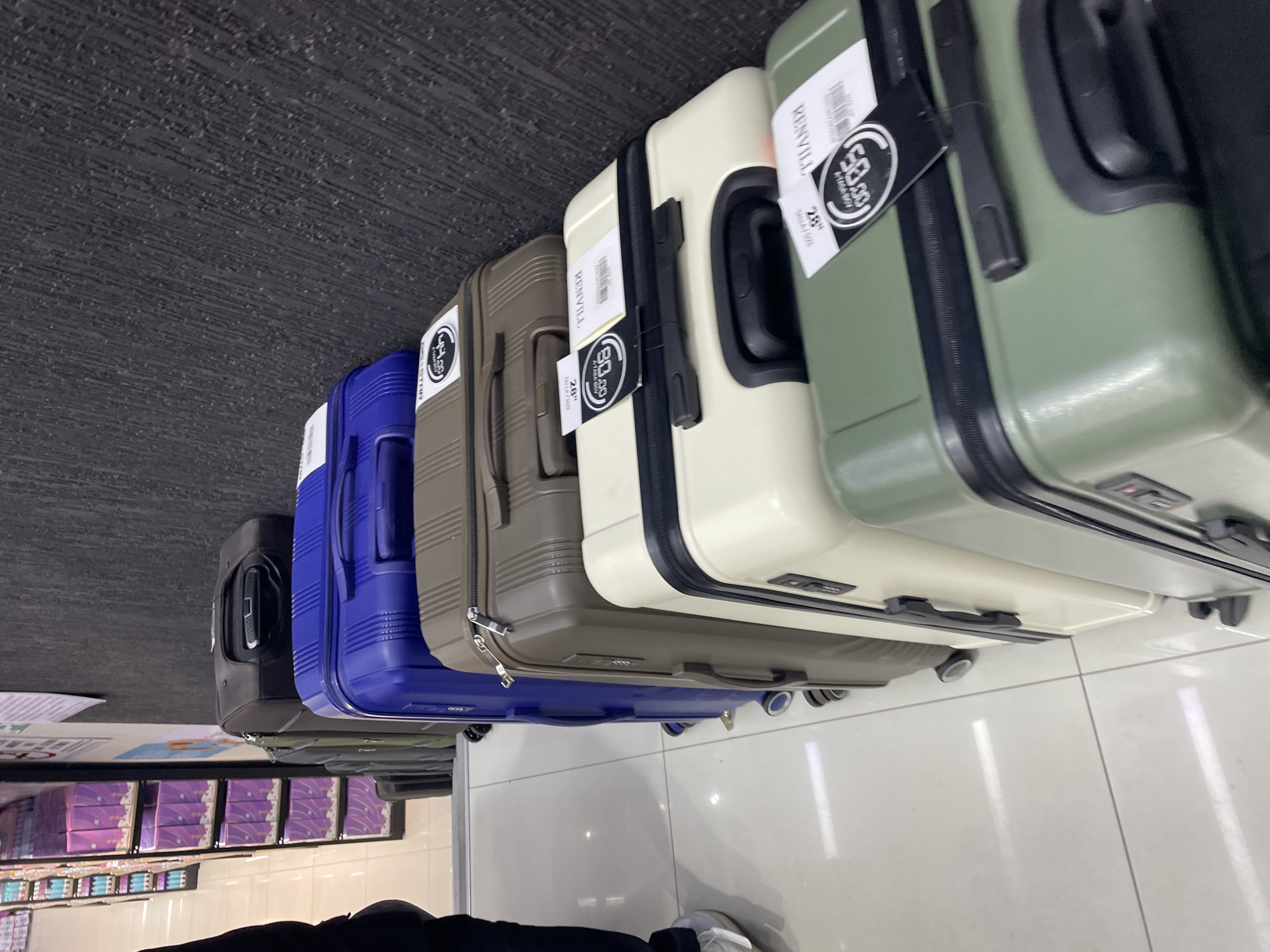 In an indoor retail environment, a row of stacked suitcases is displayed against a textured dark grey wall and a light-colored tiled floor. The suitcases are arranged in a diagonal line, with the closest one being a green, hardshell suitcase. Moving upwards and to the left, subsequent suitcases include a cream-colored hardshell suitcase, a brown hardshell suitcase, a vibrant blue hardshell suitcase, and a dark grey soft-sided suitcase. Each hardshell suitcase has black trim and handles. Price tags with the value "$59.99" are visible on some of the suitcases, along with branding that appears to read "KENVILL". To the far left, a person's legs and lower torso are visible, wearing dark pants and light-colored athletic shoes. In the background, shelves filled with products, possibly feminine hygiene items in purple packaging, are visible. The lighting suggests a brightly lit commercial space.