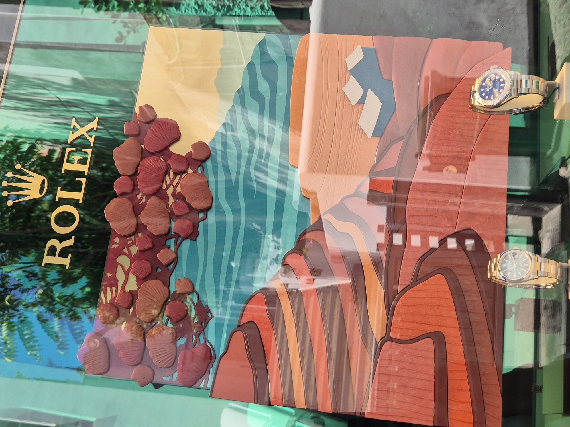 The image shows a close-up of a Rolex store window display. The display features a vibrant, layered artwork depicting a stylized landscape of terraced hills in earthy tones of red, orange, and brown, with a teal, wavy ocean or sky in the background. Above the landscape, there are decorative elements resembling red and pink shells or abstract shapes. The prominent "ROLEX" logo in gold lettering is visible at the top of the display, superimposed over a background that appears to be a photograph of lush green foliage. Two Rolex watches are displayed on pedestals in the foreground, one gold and the other a combination of gold and steel with a blue face. The watches are positioned to the left and right of the lower part of the artwork. The overall impression is of a visually rich and luxurious retail presentation.