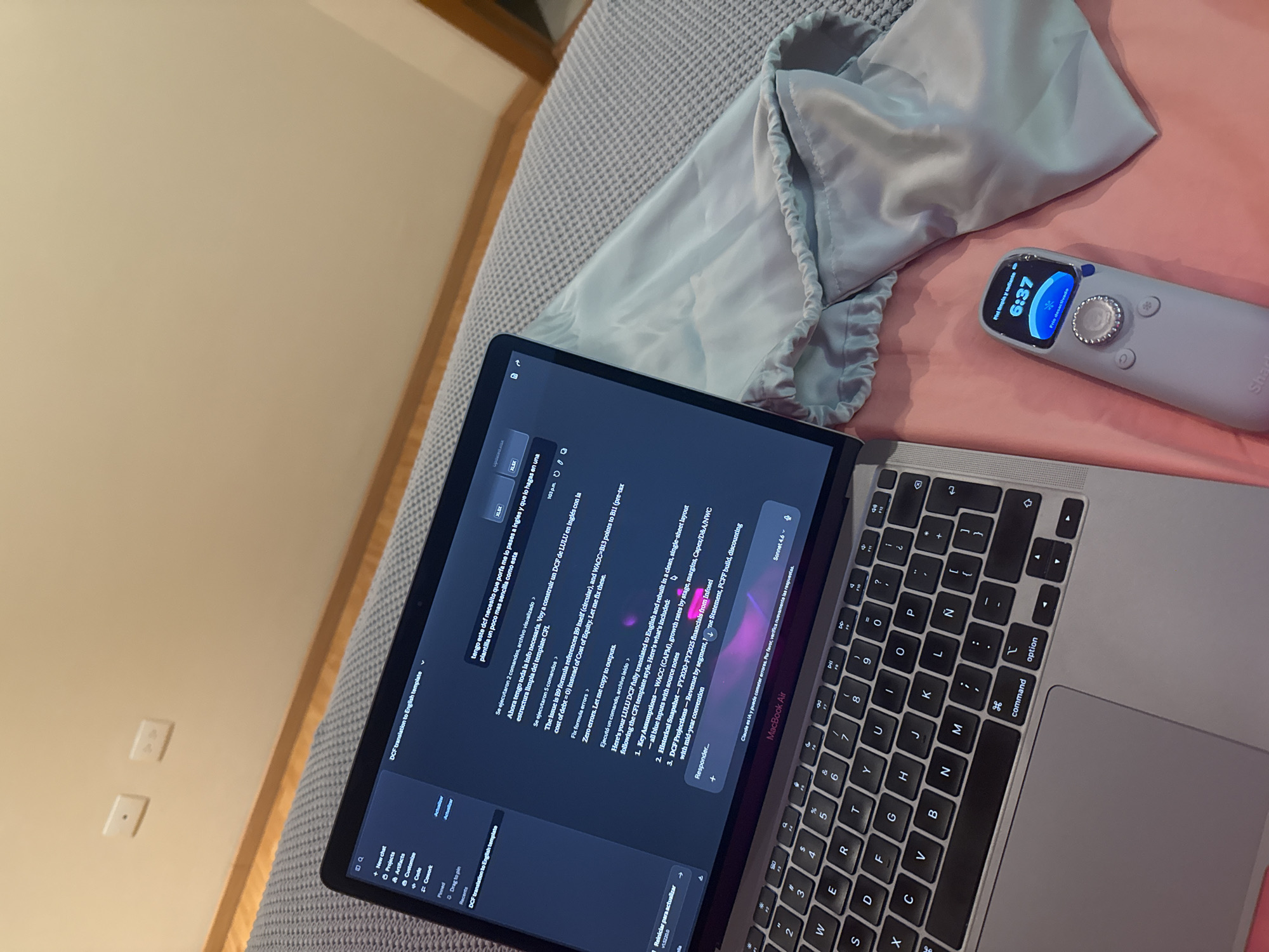 An overhead shot captures a MacBook Air laptop, a light blue satin-like bag, and an electronic device resting on a patterned grey blanket, partially covering a pink sheet. The setting appears to be indoors, likely a bedroom, with a beige wall featuring two white electrical outlets and a wooden edge visible in the background.
The MacBook Air is open, displaying a screen that shows a chat interface, likely an AI assistant. The conversation revolves around translating a Discounted Cash Flow (DCF) model to English and fixing formula errors. Text on the screen indicates questions and responses in both Spanish and English, mentioning "LULU DCF," "CFI template," "WACC (CAPM)," and "Sonnet 4.6," a reference to an AI model. A menu on the left side of the screen shows options like "New chat," "Projects," and "Artifacts." The time "103pm" is visible on the laptop screen.
To the right of the bag, a light grey elongated electronic device with a small illuminated screen is visible. The screen displays "6:37," a star icon, a water drop icon, and text in Spanish: "Piel limpia y radiante" (Clean and radiant skin) and "Frío desactivado" (Cold deactivated). Part of a brand name, "Sha," is visible on the device's body. The combination of the laptop for work/study and the skincare device suggests a moment of relaxation or personal care alongside digital activity in San Bernardino Tlaxcalancingo, Mexico.