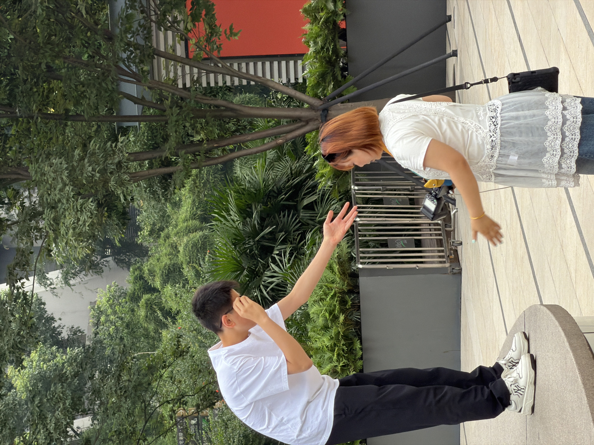 In Yuzhong District, China, two individuals are interacting outdoors. A young man, wearing a white t-shirt and black pants, is leaning forward with his right hand extended, palm open, towards a woman. He is wearing white sneakers with black accents. The woman, facing away from the camera, has reddish-brown hair and is wearing a white lace-trimmed top and blue jeans. She is holding a black camera with a yellow strap in her left hand and also has her right hand extended, palm open, towards the man, suggesting an exchange or greeting. They are standing on a paved area with a circular raised platform in the foreground. In the background, there are lush green trees and foliage, a metal railing, and a modern building with an orange section.