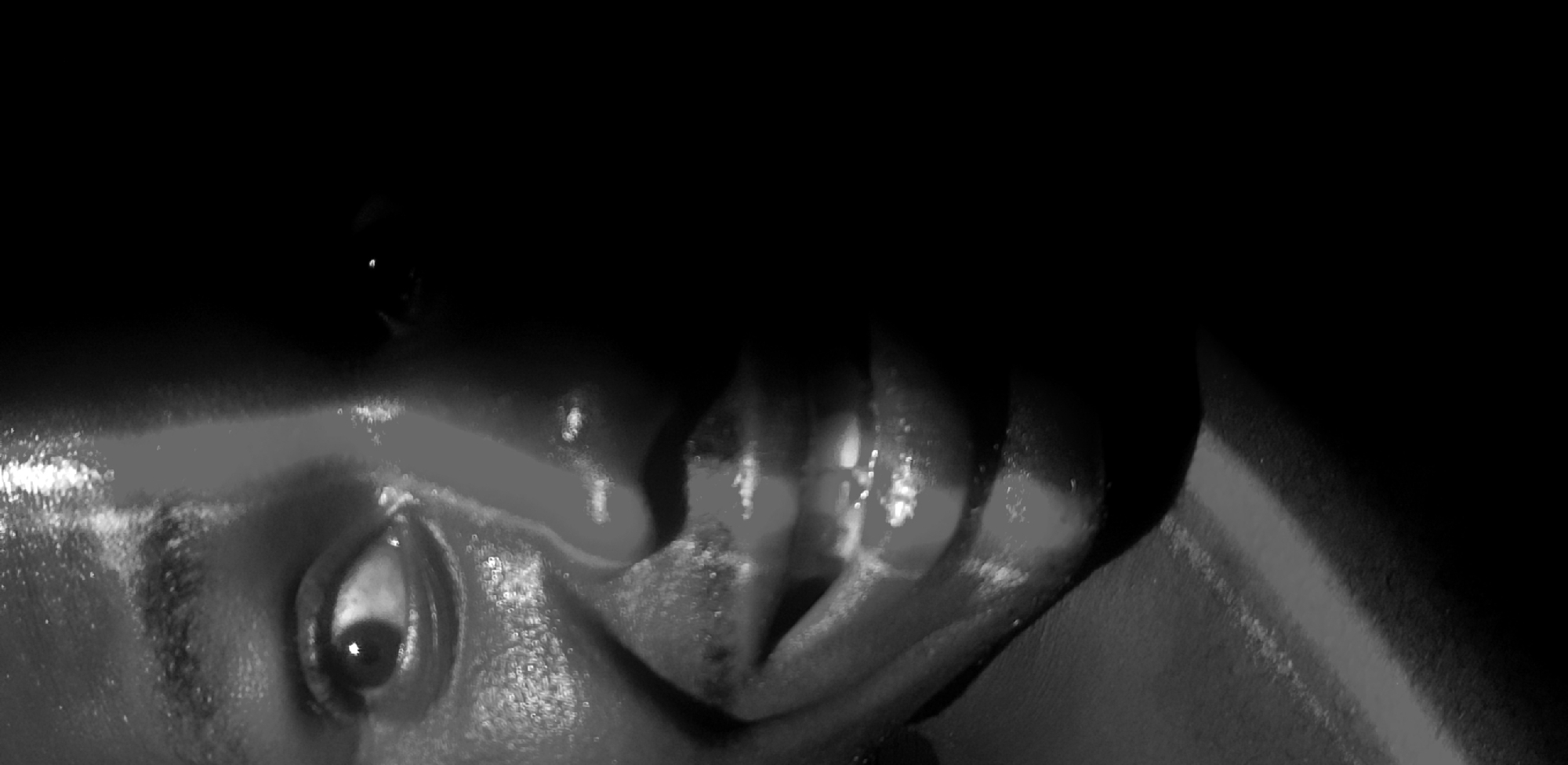 A close-up, black and white, low-angle shot captures the left side of a person's face, with the right side obscured by deep shadow. The subject's left eye is visible, looking slightly to the left, with a glint of light reflecting in the pupil. The cheekbone and forehead are illuminated, highlighting a shiny, possibly sweaty, texture. The mouth is open in a smile, revealing teeth and glistening lips, with the lower lip appearing slightly fuller. A portion of the neck and shoulder is visible on the lower left, with a textured appearance. The background is entirely black, creating a stark contrast with the illuminated facial features. The image is devoid of any contextual information about its location in Narayi, Nigeria, or any specific details pertaining to a city environment.
