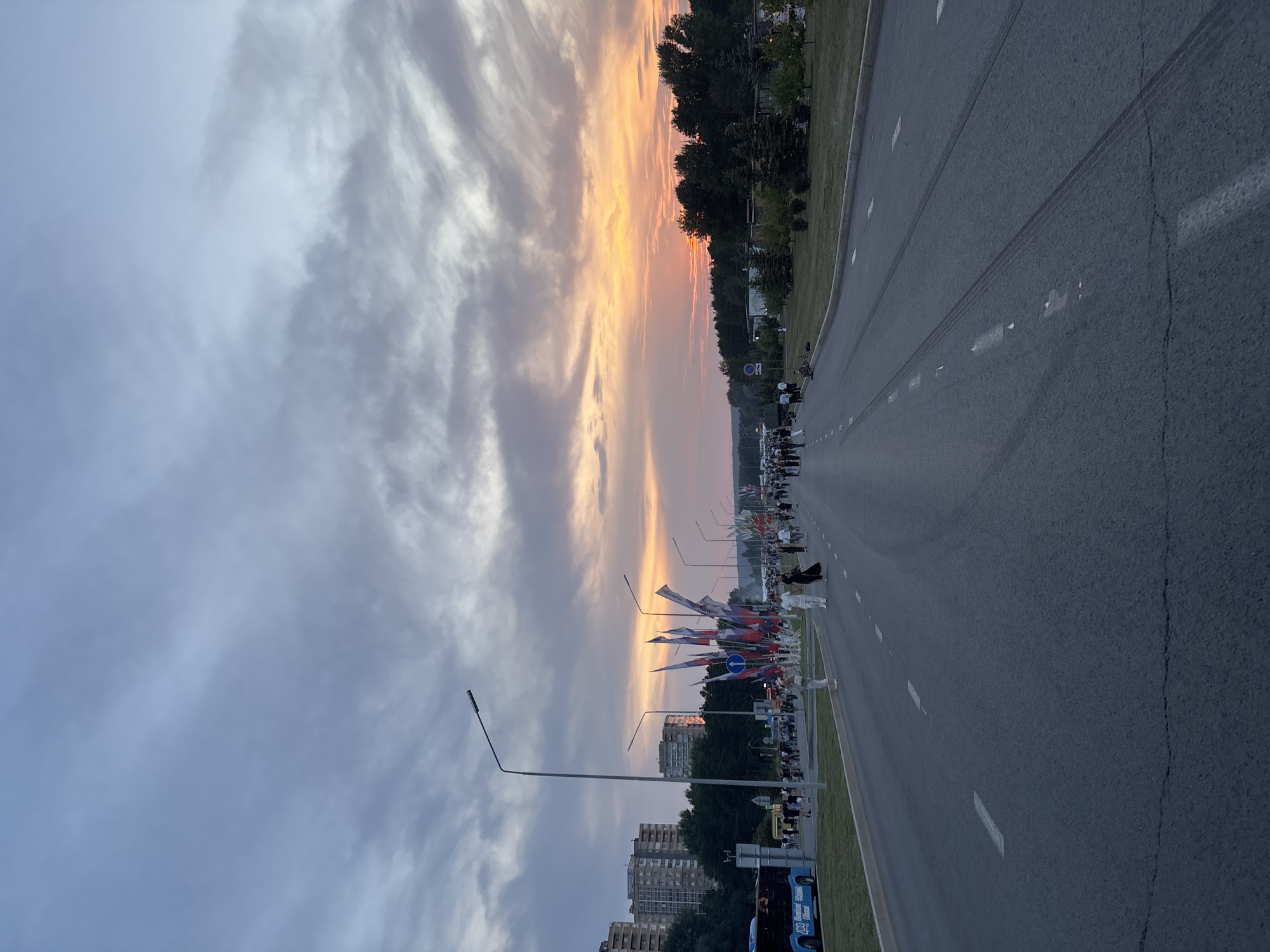 The image depicts an outdoor scene during dusk on a wide asphalt road. The sky is filled with dramatic clouds, transitioning from a soft grey at the top to vibrant oranges and pinks near the horizon, indicating a sunset. Along the right side of the road, a line of evergreen trees borders a grassy area. On the left side, a blue bus is parked, and beyond it, several multi-story buildings are visible.
The road is marked with white lane lines and appears to be a significant thoroughfare. A procession of people is walking along the right side of the road, some in white attire, others in darker clothing. Many flags, appearing to be national or ceremonial flags, are displayed on poles along the right side of the road, interspersed with streetlights. The overall atmosphere suggests a public event or gathering.
The specific location is identified as the city of Naberezhnye Chelny, Russia. The scene captures a moment of twilight with a noticeable presence of a public procession and decorative flags, set against an urban backdrop with residential buildings.