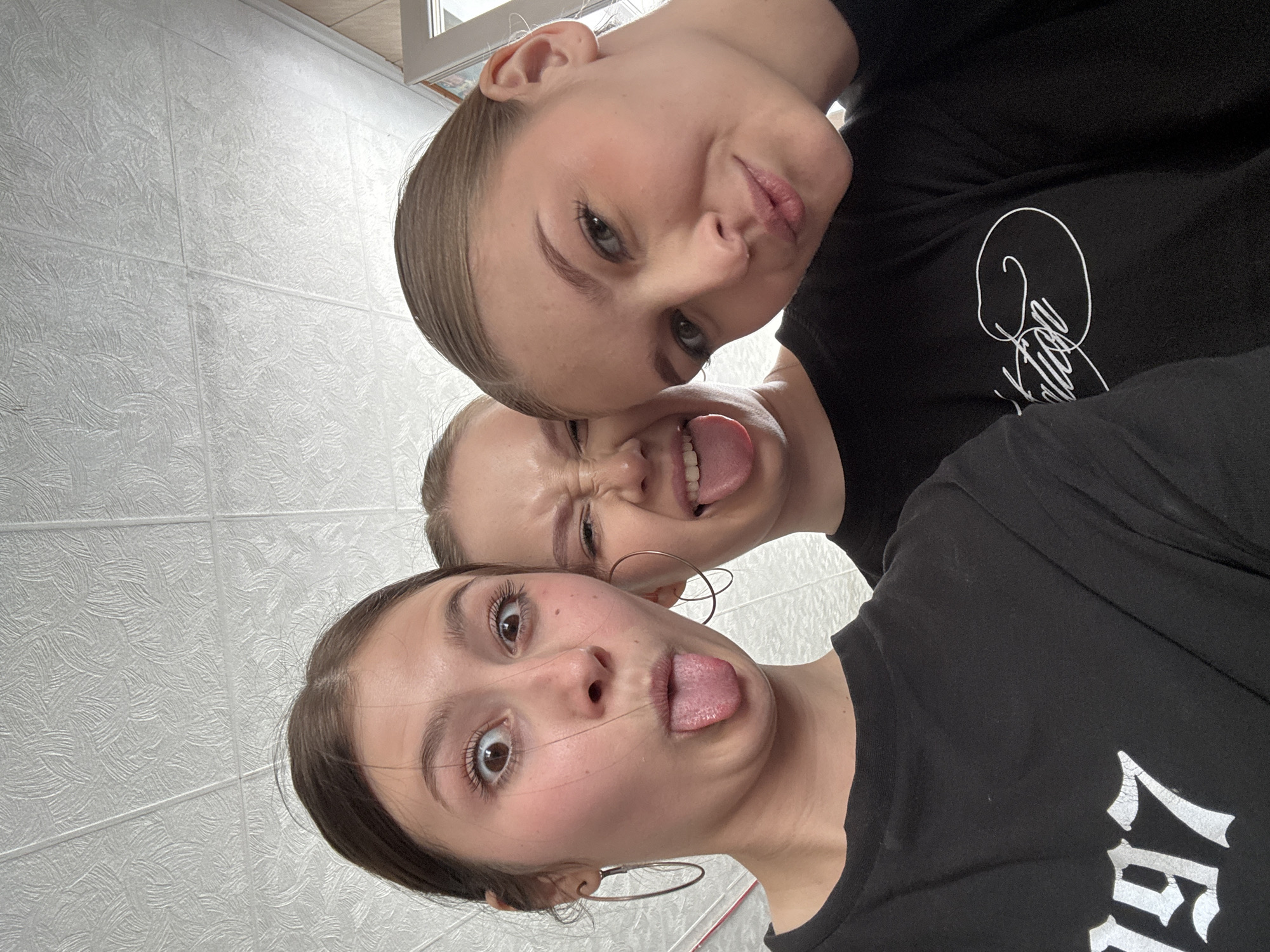 Three young women are posed closely together, presumably taking a selfie. They are all wearing black t-shirts. The top woman is making a kissing face. The woman in the middle is sticking her tongue out and squinting. The woman in the foreground is also sticking her tongue out and widening her eyes. The background is a light-colored wall with a textured pattern. A partial window frame is visible in the top left corner. No features indicating Chaikovsky are visible.