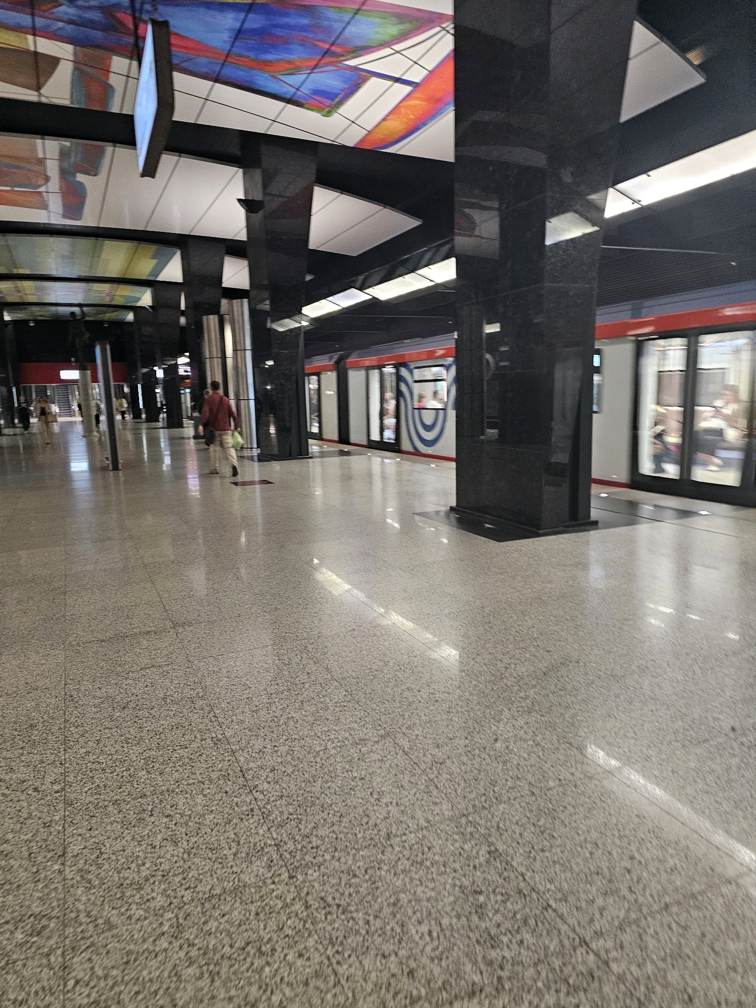 The image depicts a modern subway station. In the foreground, a man in a red shirt and light-colored pants is walking away from the camera, carrying a bag. He is on a spacious, polished floor with a speckled grey and white pattern. Several large, dark, rectangular pillars line the platform, some with reflective surfaces. Above, a colorful abstract mural is visible on the ceiling. To the right, a sleek, modern train with red trim and large windows is stopped at the platform. Passengers are visible inside the train, some looking out the windows. The station lighting is bright and overhead. The overall impression is one of a clean, contemporary public transportation hub.