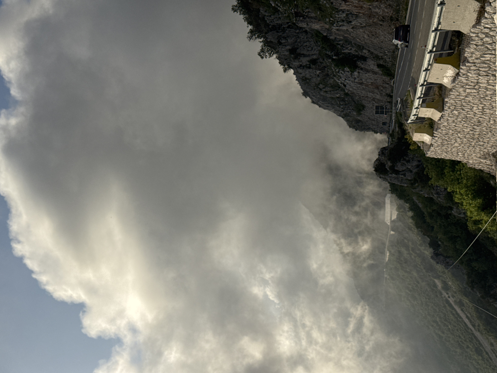 The image shows a road built along a cliff in Brajici, Montenegro. The cliff is rocky with some vegetation. A stone wall and metal guardrail border the road. A car is visible on the road. The sky is filled with heavy clouds. Fog or mist hangs over the landscape below the road.