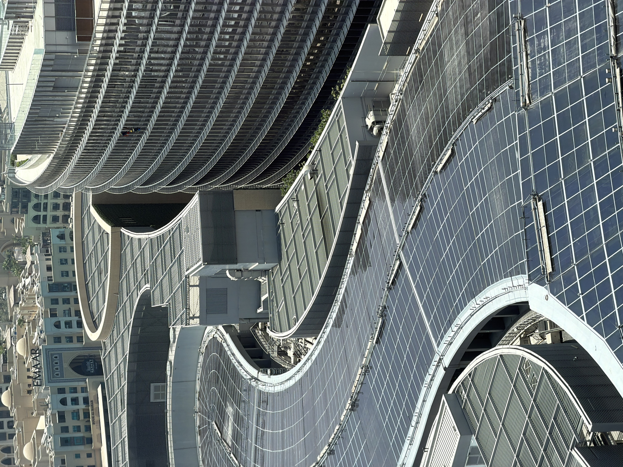 The image shows an aerial view of the Dubai Mall with the Burj Khalifa in the background. The mall features a large, curved roof covered in solar panels. The building is a modern architectural design, with clean lines and a metallic facade. In the distance, the iconic Burj Khalifa skyscraper can be seen. The sky is clear and blue, indicating a sunny day. The scene is set in Dubai, United Arab Emirates.