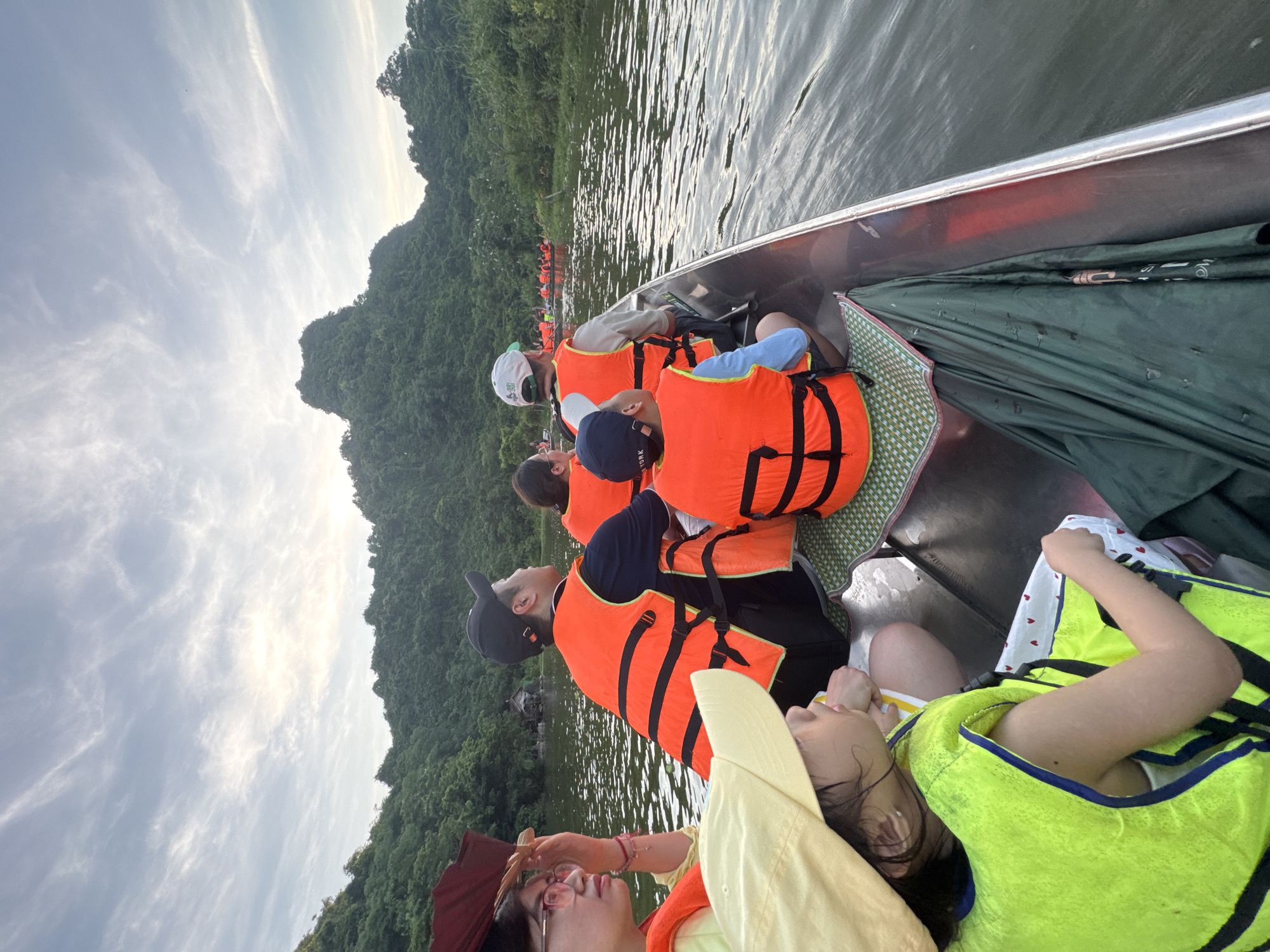 The image shows a group of people in a boat, likely on a river or lake, with lush green hills in the background. The sky is partly cloudy. The people are wearing orange and yellow life vests, indicating they are engaged in a water activity. In the foreground, a young child with a yellow hat and vest is facing away from the camera. Behind them, adults and other children are also seated in the boat, some looking forward and others to the side. The boat appears to be made of metal. In the distance, other boats are visible, suggesting a tour or group outing. The overall setting suggests a scenic natural environment, possibly a tourist destination. The location in Yên Sơn Ward, Vietnam, aligns with such a landscape, known for its natural beauty and waterways.