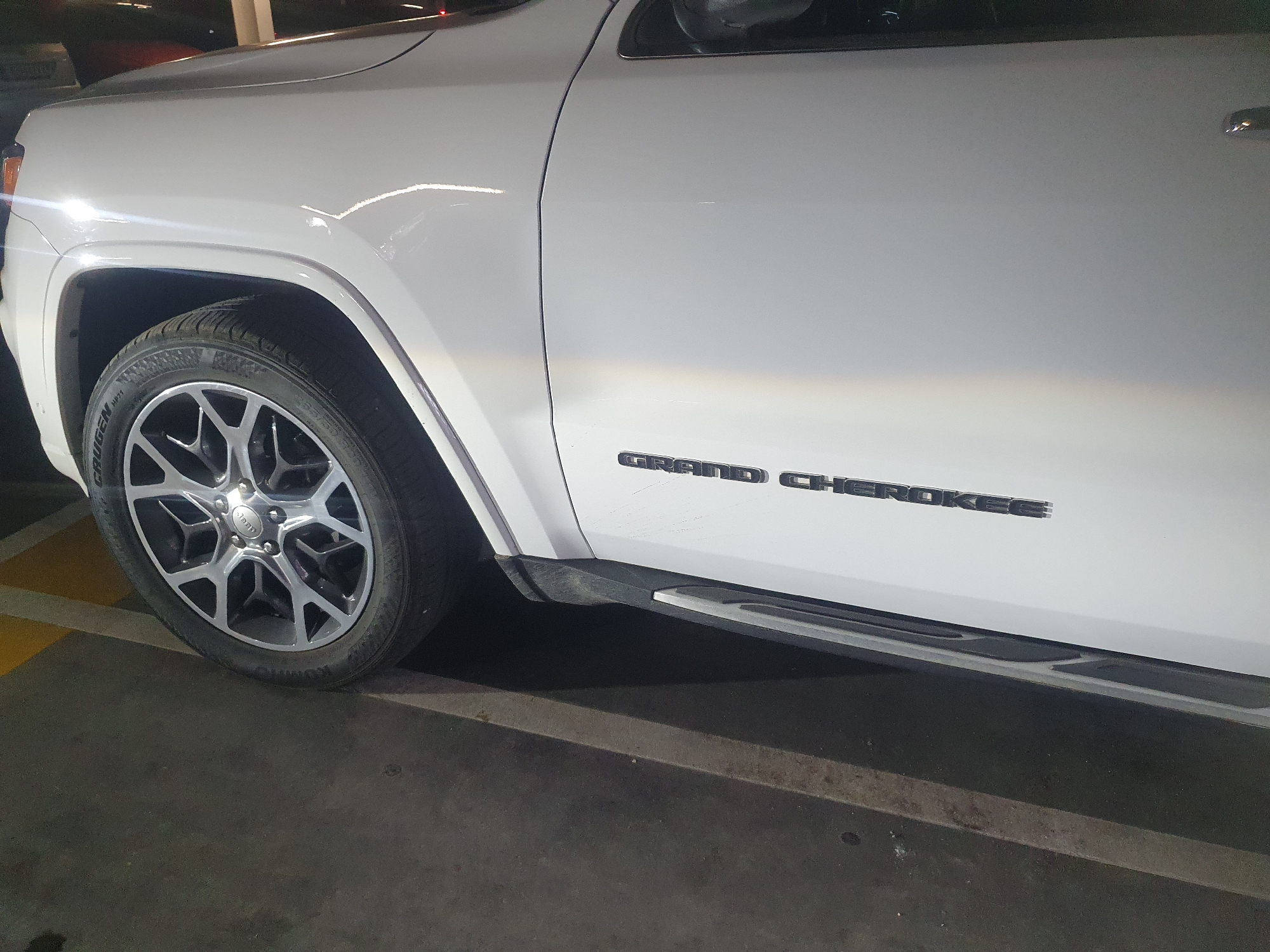 The image shows the front driver's side of a white Jeep Grand Cherokee parked on a concrete surface with yellow parking lines. The tire is visible and has a silver and black rim. The words "GRAND CHEROKEE" are displayed in black lettering on the side of the vehicle. The scene appears to be indoors, likely in a parking garage. There are no visible people in the image. The location in Plovdiv, Bulgaria, is not apparent from the given visual information.