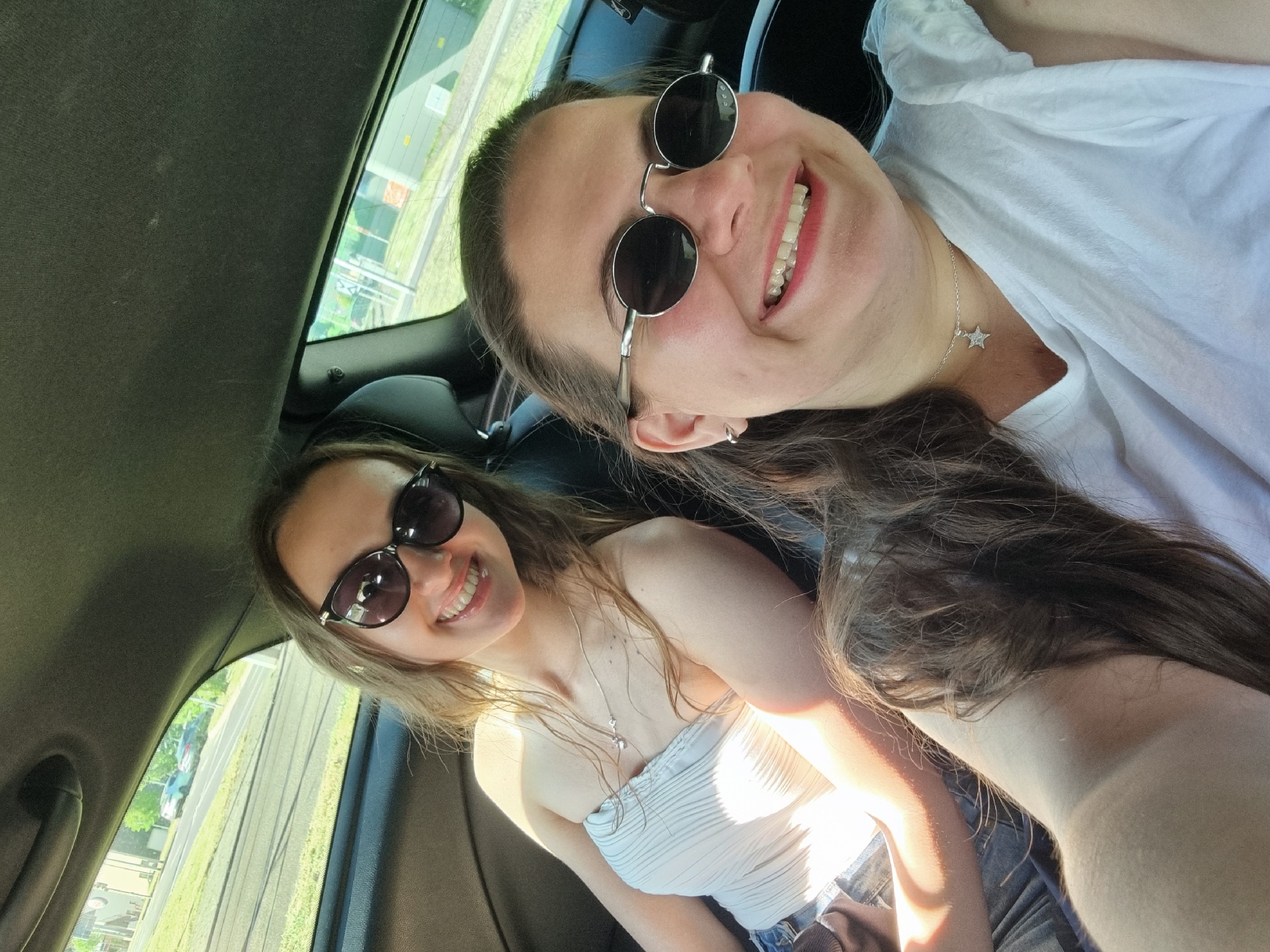 Two young women are in a car. One woman is in the front passenger seat, wearing a white strapless top, sunglasses, and a necklace. The other woman is behind her, wearing a white shirt, sunglasses, and a necklace with a star pendant. Both women are smiling.
Outside the car window are tram tracks, indicating they are likely in a city. Given the location is Saarbrücken, Germany, these are likely the tracks for the Saarbahn. There are grassy areas and some buildings visible outside the window.