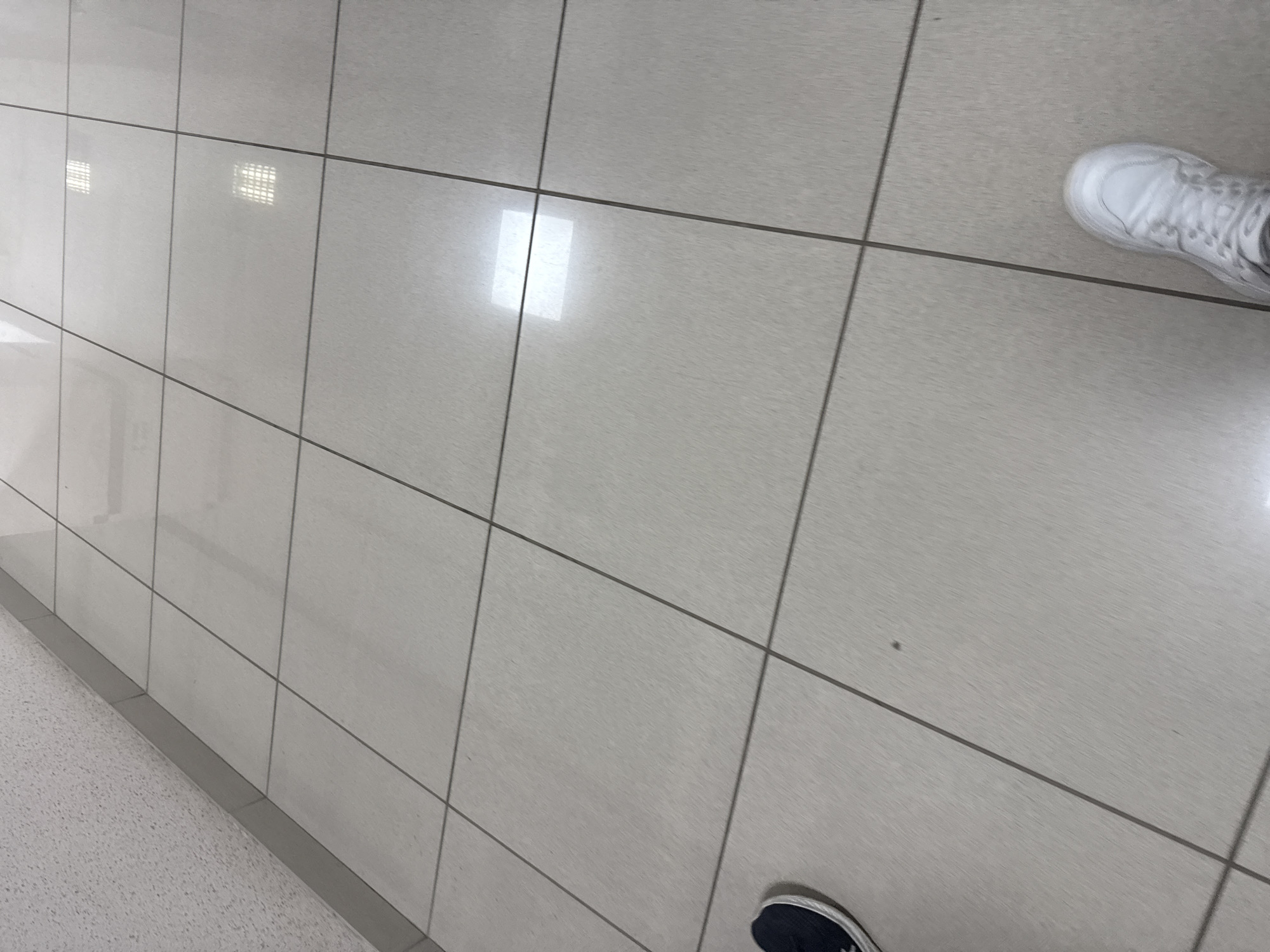 The image shows a tiled floor in what appears to be an indoor corridor. The tiles are large, square, and light-colored, possibly beige or light gray, with dark grout lines. There are reflections on the floor, indicating it is clean and possibly polished. In the upper left, a textured wall with a baseboard can be seen. The scene is observed from a first-person perspective, with two feet visible: one wearing a dark sneaker on the left side of the frame, and another wearing a white sneaker on the right side. The white sneaker is closer to the viewer and in focus. The overall impression is of a clean, modern, and possibly institutional or commercial interior. There is no specific indication of the city of Perm, Russia, or any other location.