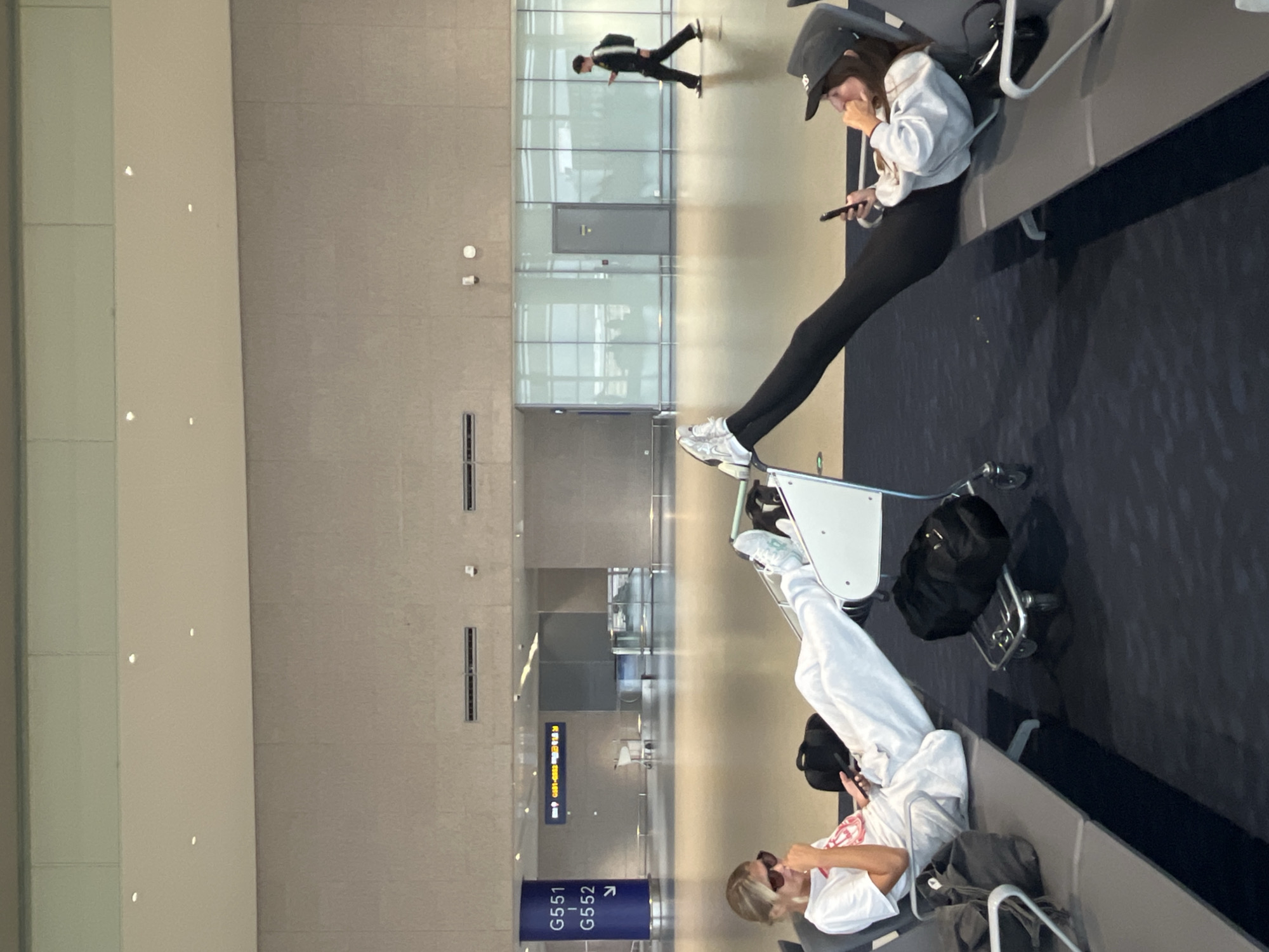 In Pudong, China, two young women are seated on airport lounge chairs, facing each other with their legs extended and resting on luggage carts. The woman on the left, wearing sunglasses, a white t-shirt, and light-colored sweatpants, holds a phone in her hand. The woman on the right, sporting a baseball cap, a light grey hoodie, and black leggings, also holds a phone. They are positioned in a spacious airport terminal with large windows, visible seating arrangements, and a distant figure walking away from the camera. A dark-colored carry-on bag rests on the floor next to the woman on the left, and a black duffel bag is on the luggage cart between the two women. Overhead lighting illuminates the terminal, and signs indicating gate numbers are visible on a blue pillar in the background. The overall scene depicts a moment of waiting at an airport.