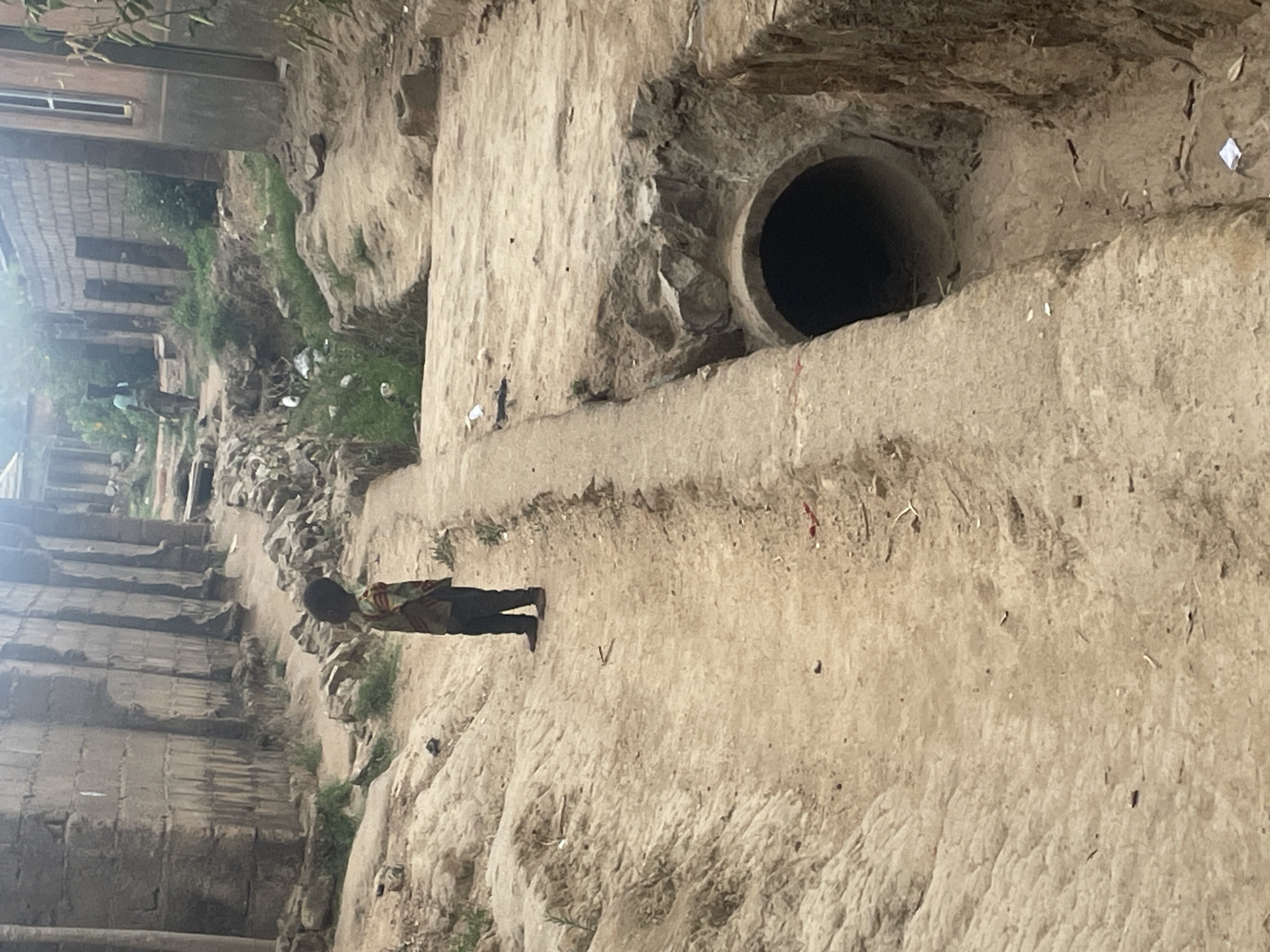 The image depicts an outdoor scene in a semi-urban or rural setting, characterized by unpaved, uneven terrain. A wide, unpaved channel or path, composed of dirt and sand, traverses the scene, showing signs of erosion and rough texture.
In the mid-ground, a lone individual, likely a child or young person, stands on this earthy path. They are wearing a striped or patterned long-sleeved shirt and dark trousers, with their back partially turned, suggesting they are observing the surroundings or moving slowly.
A prominent feature on the right side of the frame is a large, dark circular opening of a concrete pipe or culvert, embedded within an earthen bank. Its interior is obscured by darkness, indicating a deep conduit. The ground around this structure is rough, appearing to be compacted earth and rock.
In the background, on higher ground to the left, several structures resembling buildings are visible. They are constructed from light-colored blocks or concrete, with one building featuring a distinct rectangular window. Sparse patches of green vegetation, including grass and small shrubs, are scattered along the earthen banks and within the channel. Various pieces of debris, such as white and dark fragments, are visible on the ground, suggesting litter.
The lighting indicates daytime, possibly morning or afternoon, with natural light illuminating the scene evenly. The ground appears dry, suggesting dry weather. The overall impression is of an ordinary, rugged environment. No specific activity beyond the presence of the individual is discernible. There is no visible text or explicit emotional context. The setting aligns with the provided context of Jos, Nigeria, reflecting typical infrastructure and environment in such areas.