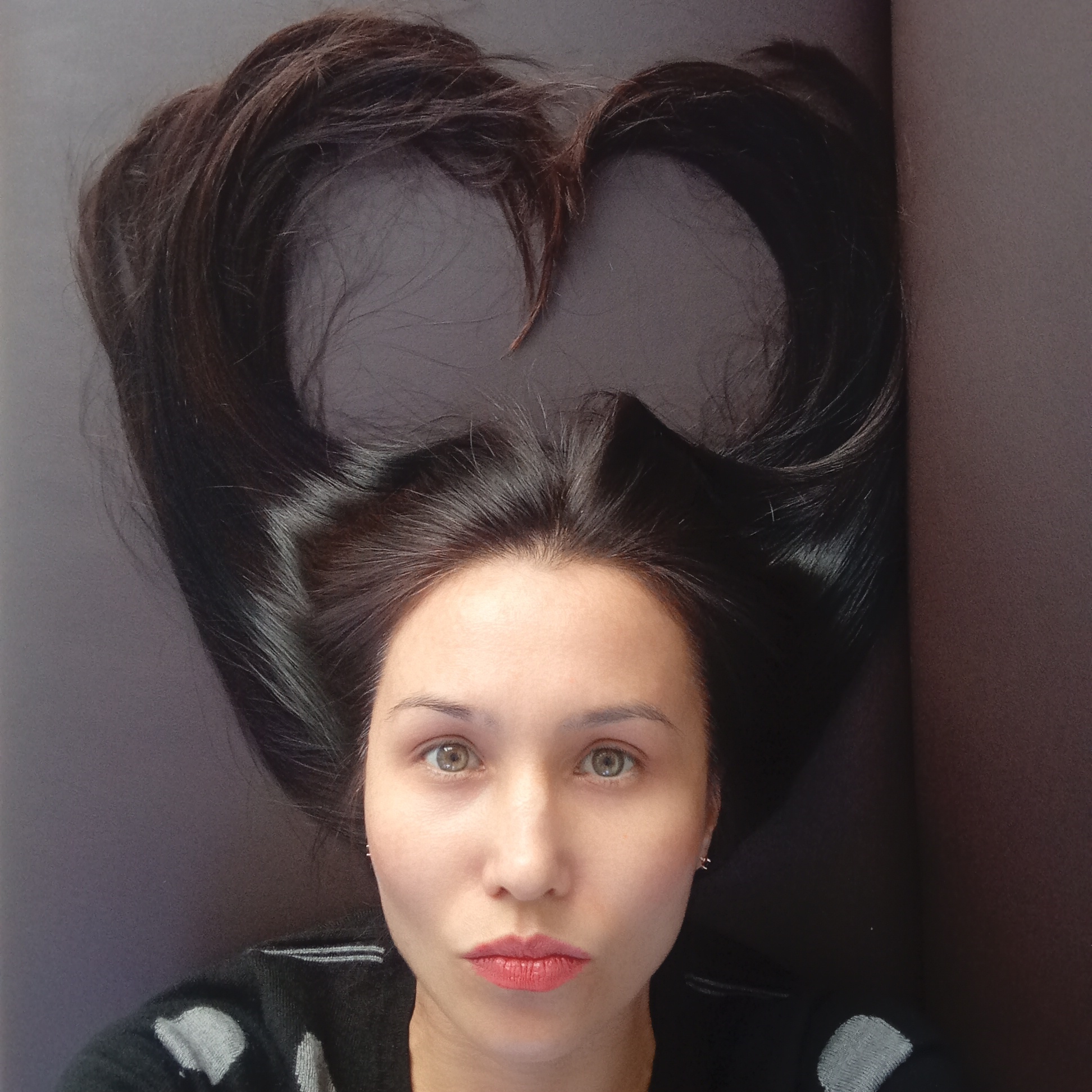 A portrait photograph captures a young woman from the chest up, positioned against a dark, plain background. Her long, dark brown hair is artfully arranged above her head, forming a prominent heart shape. She has light skin, large, light greenish-hazel eyes, and dark eyebrows, looking directly at the viewer with a neutral expression and slightly pursed lips colored with reddish-pink lipstick. Small silver stud earrings are visible on her earlobes. She wears a dark sweater with light grey patterns visible on her shoulders. The background is a solid, dark color, possibly a textile or paper, and shows a slight crease or fold on the right side. The image showcases a creative pose, focusing on the woman's face and the heart-shaped hair arrangement. No text is visible in the image. The location context for this media file is Izhevsk, Russia.