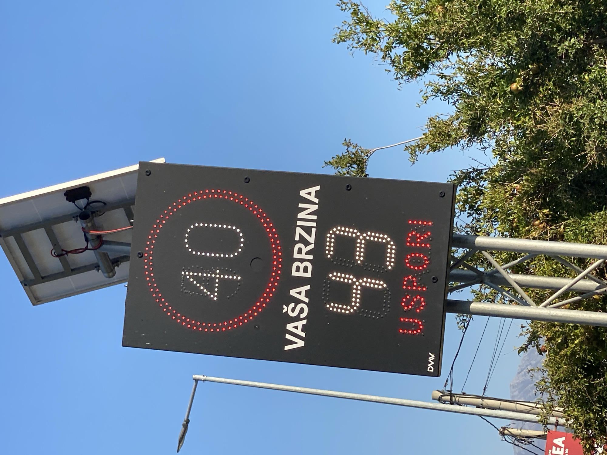 A roadside speed display sign is mounted on a metal pole under a clear blue sky. The sign is rectangular with a black background. At the top, a red circle encloses the white LED display of the number "40". Below this, in white LED text, is "VAŠA BRZINA", which translates to "YOUR SPEED". In the center, a larger white LED display shows the number "43". Below this, in red LED text, is "USPORI", which translates to "SLOW DOWN". A small "DMV" logo is visible in white LED text in the lower left corner of the sign. The sign is powered by a solar panel mounted on top of the pole. To the left of the sign is a street light. In the background, there is a mountain range. The foreground shows green foliage and some small, round, reddish fruits on a tree. The overall scene appears to be on the outskirts of Zgrade, Montenegro, likely along a road.