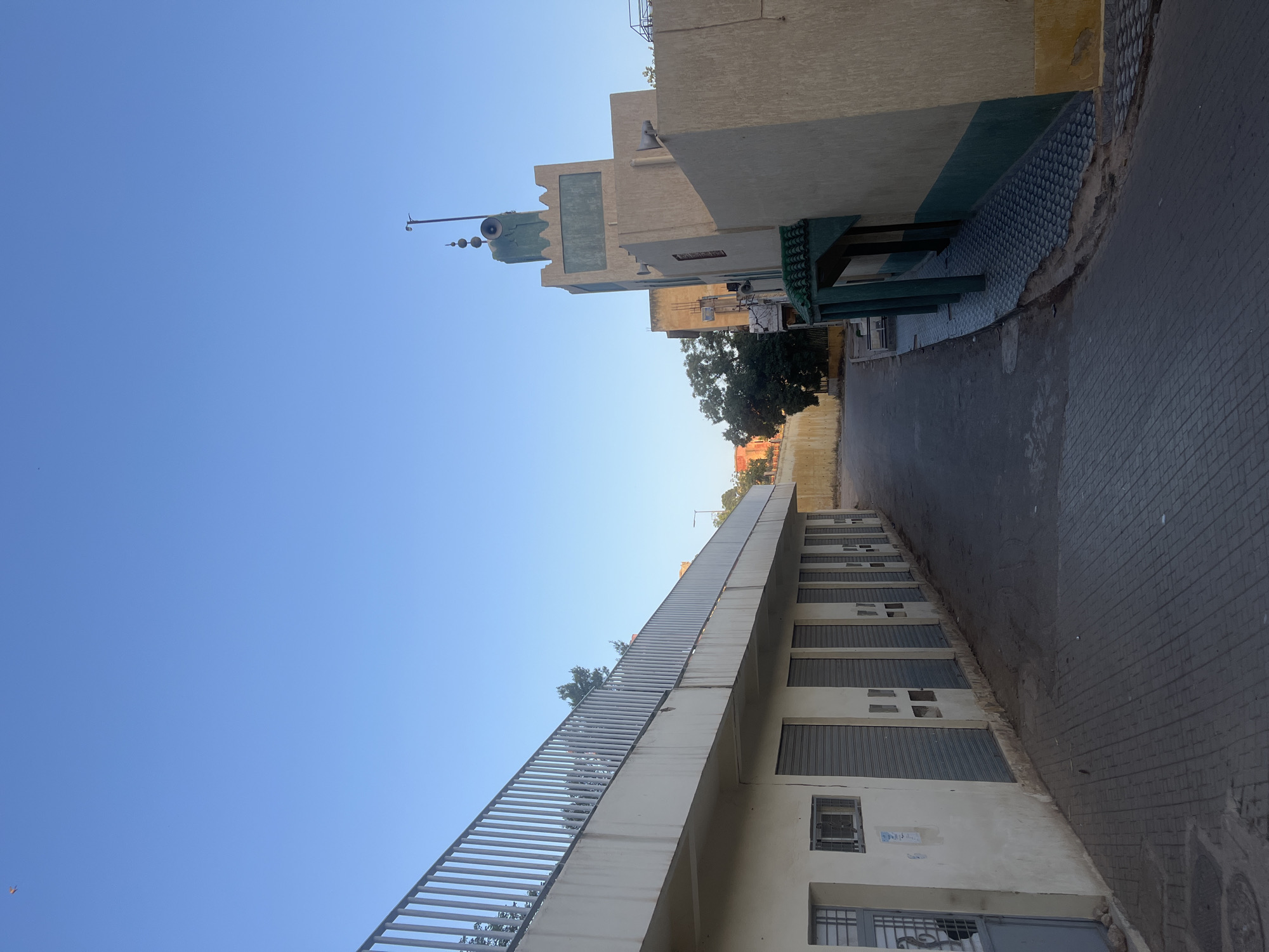 The image depicts a street scene in El Hajeb, Morocco, under a clear blue sky. On the left, a two-story building features a concrete facade with a metal railing on the upper level. The lower level has several closed roll-up doors and a barred window. The ground is paved with gray interlocking bricks. On the right, a tan building with green accents, resembling a mosque, is visible. It has a minaret topped with speakers. Trees and a yellow wall are present in the background. The street itself is paved with asphalt.