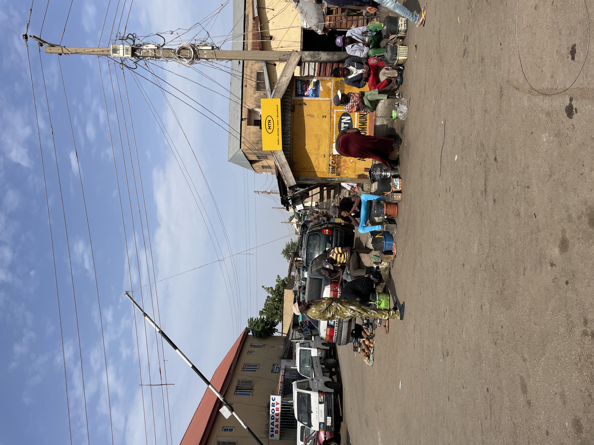 This media file captures a vibrant street scene in Jos, Nigeria, during the daytime under a clear blue sky with scattered white clouds. The setting appears to be a busy commercial area with buildings lining a paved road.
Numerous people are present, engaged in various activities. Many individuals are seated on the ground or low stools alongside the road, often surrounded by various items, suggesting street vending or waiting. These items include what appear to be small appliances, electronics, and a pile of coconuts. Other people are seen standing or moving along the road. Several vehicles, including cars and small vans, are parked along the street.
Prominent elements include a tall wooden utility pole on the left, heavily laden with a dense network of electrical wires and components. Further down the street, a black and white striped barrier arm, possibly a checkpoint or entrance gate, is visible. Buildings on both sides of the street feature signs. A bright yellow sign on a building reads "MTN MoMo • Airtime • Data • Broadband." Below it, another partially visible sign mentions "SIM CAR REGISTRATION CE" and "COMMUN." Further along, a building displays the name "SHADORC BAKERY." A poster with "EVERYBODY" is also partially visible on a building facade. The road itself is asphalt, showing some wear, with minor debris such as a plastic bottle and a metallic hoop visible on its surface. The overall impression is a bustling urban environment characterized by informal commerce and daily activity.