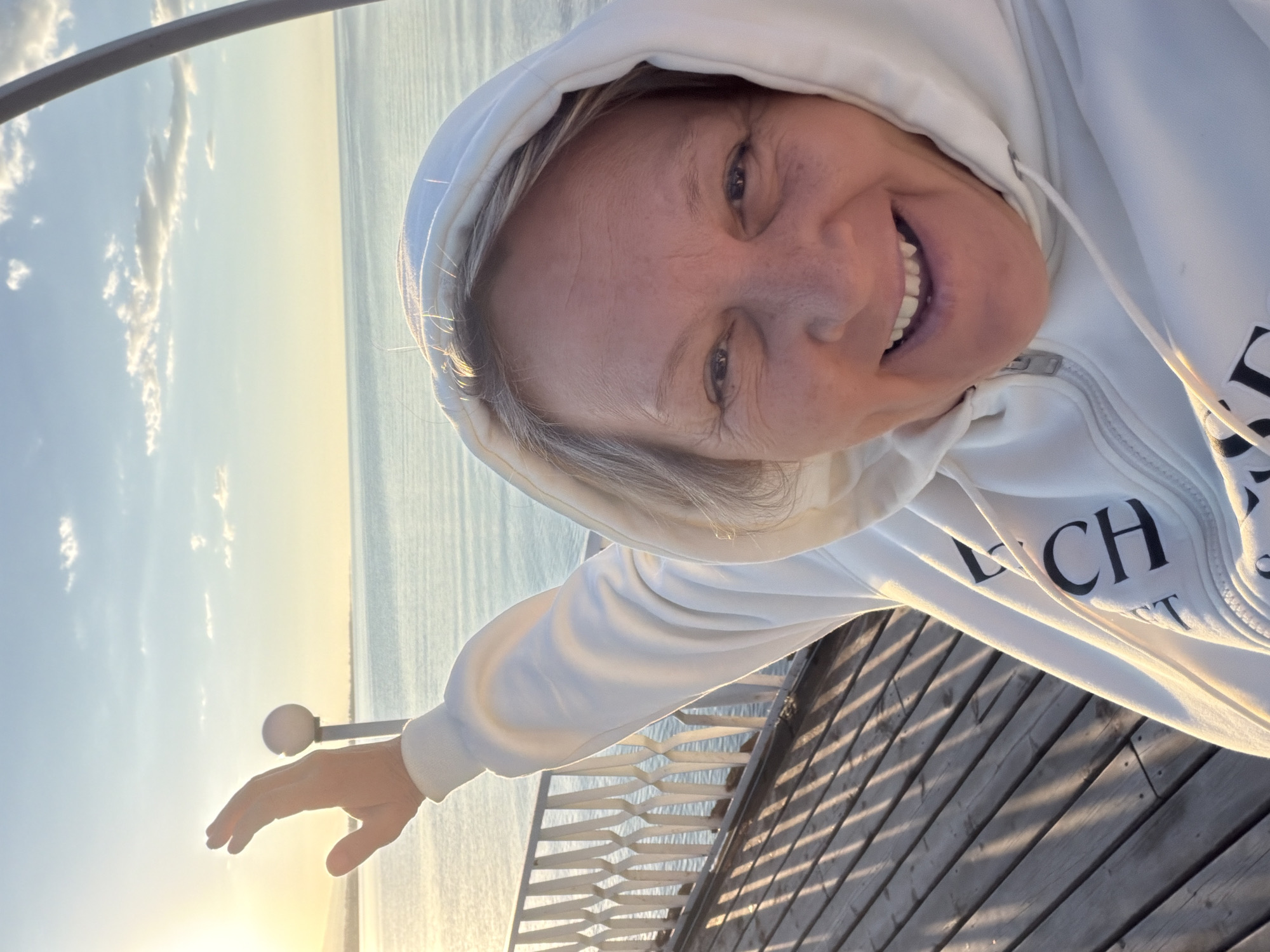 A woman with short, graying hair, wearing a white hooded sweatshirt, smiles at the camera. Her right arm is raised, and her hand is open with fingers spread. She is standing on a wooden pier, indicated by the planks and railings visible in the foreground and middle ground. The background shows a calm body of water, likely a lake or sea, reflecting the sky. The sky is a mix of blue and white clouds, with the sun setting or rising on the left side of the frame, casting a warm glow and long shadows across the pier. A lamppost with a spherical light fixture is positioned behind her raised arm.