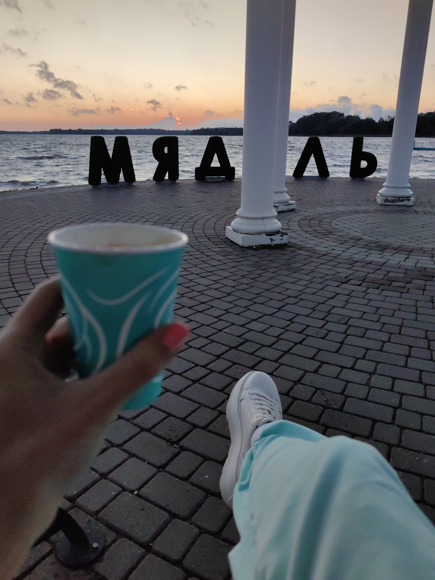 In Myadzyel, Belarus, a person's hand, with a pink fingernail, holds a turquoise paper cup with a white lid, containing a beverage. The cup features a white abstract design. The person's legs are visible, clad in light blue sweatpants and white platform sneakers, resting on a paved area of interlocking gray bricks. In the background, large black letters spelling "МЯДЗЕЛЬ" are arranged in front of a body of water. The letters are positioned on the paved area near the water's edge. The sky above is a gradient of orange and pink hues, indicating dusk or dawn, with scattered clouds. Two white columns stand to the right of the letters, framing the scene. The overall atmosphere suggests a moment of relaxation by the water.