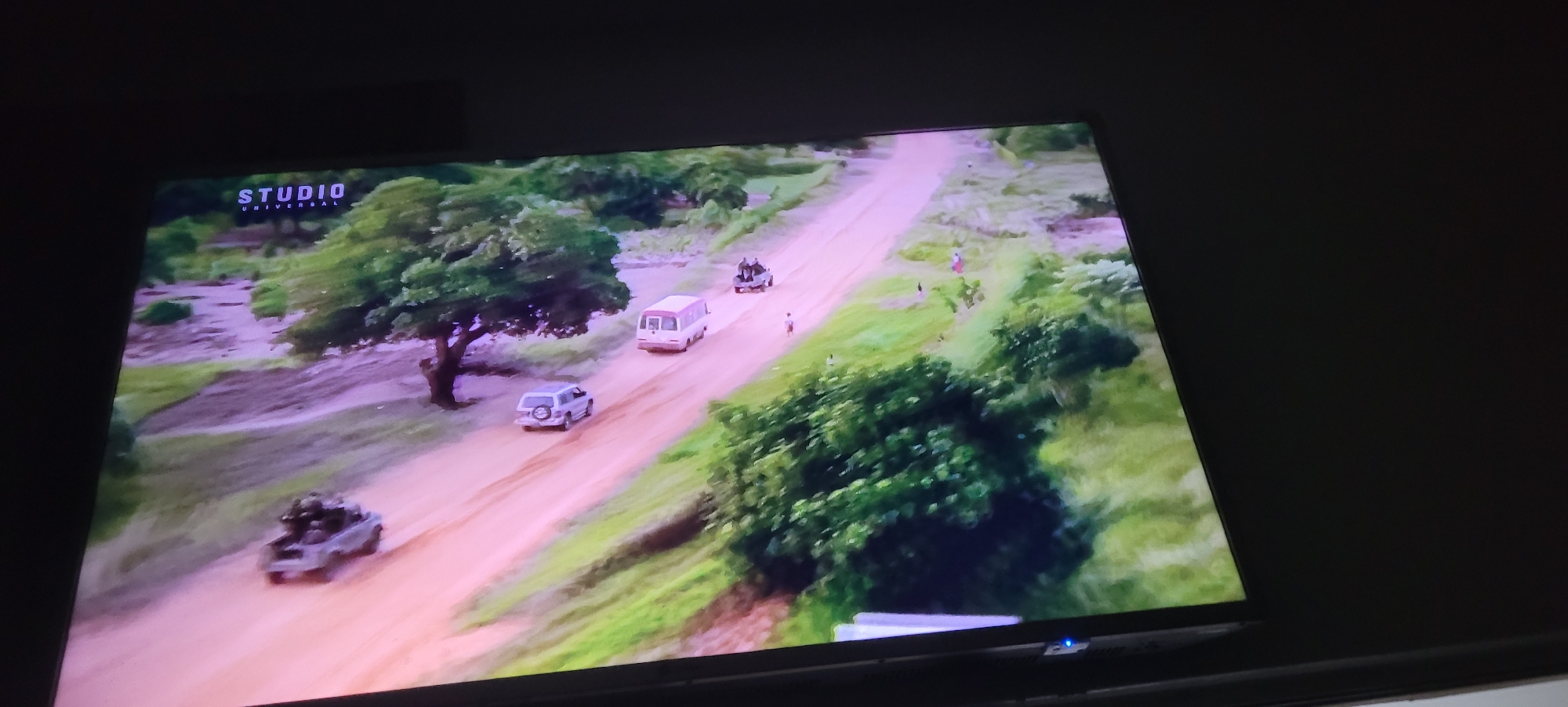 An aerial view captures a dusty red road cutting through a landscape dotted with green trees and vegetation. Several vehicles are present: a military-style jeep with people in the back is moving down the road, followed by a white bus, and then a silver SUV. In the distance, another dark colored vehicle is visible. A few individuals appear to be walking or standing near the road. The scene is brightly lit, suggesting daytime. The "STUDIO UNIVERSAL" logo is visible in the top left corner of the frame. The environment appears to be a rural or semi-rural area. There are no discernible landmarks that would definitively place this scene in Jos, Nigeria.