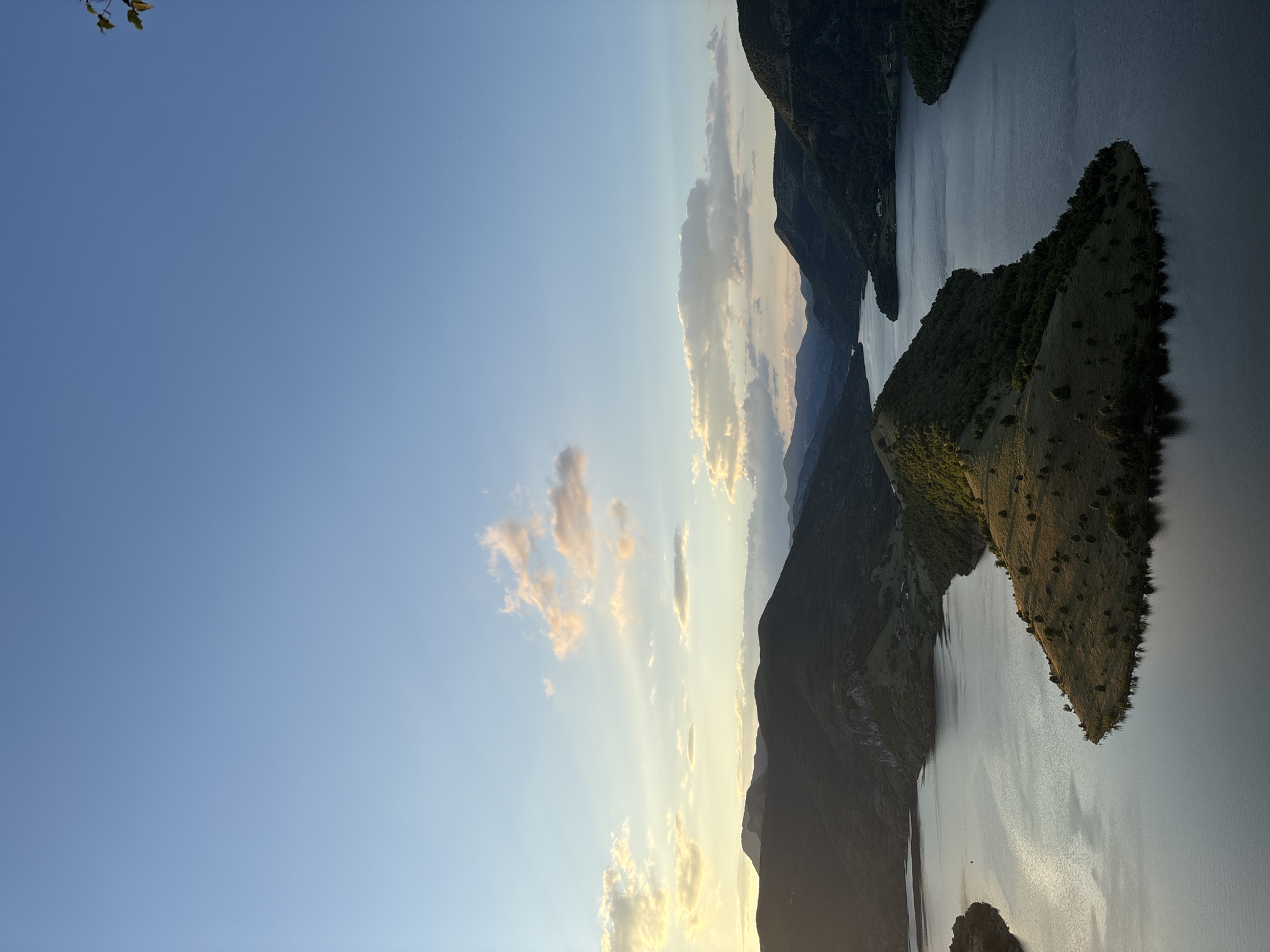 An aerial view captures the winding Ardino River as it flows through a valley surrounded by lush, tree-covered mountains. The sun is low in the sky, casting a warm glow and creating long shadows across the landscape, suggesting either sunrise or sunset. A prominent, isolated peninsula of land, covered in dry grass and sparse trees, juts out into the river, creating a distinctive "snake head" shape, a well-known landmark of the city of Borovitsa, Bulgaria. The water reflects the soft hues of the sky, with scattered clouds tinged with pink and orange. The overall scene is serene and picturesque, showcasing the natural beauty of the Bulgarian countryside.