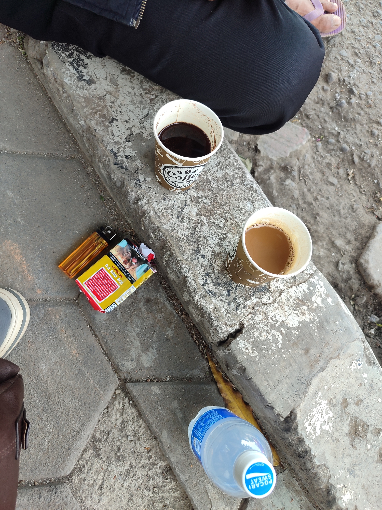 This high-angle, medium shot, captured on a paved surface, shows a still life of beverages and smoking paraphernalia. In the upper portion of the frame, a person's black pants and a purple flip-flop are visible, suggesting they are seated on a concrete ledge. Two disposable coffee cups, one with dark coffee and the other with lighter coffee, sit on the ledge. To the left of the cups, a yellow disposable lighter rests beside an open pack of "DJI SAM SOE" cigarettes, featuring a health warning in Indonesian. The ground is a textured paving of gray interlocking stones. A bottle of Pocari Sweat is positioned in the lower right corner. The scene is set outdoors, likely in a public area of Tegal, Indonesia, evidenced by the cigarette brand and the Indonesian text on the cigarette pack.