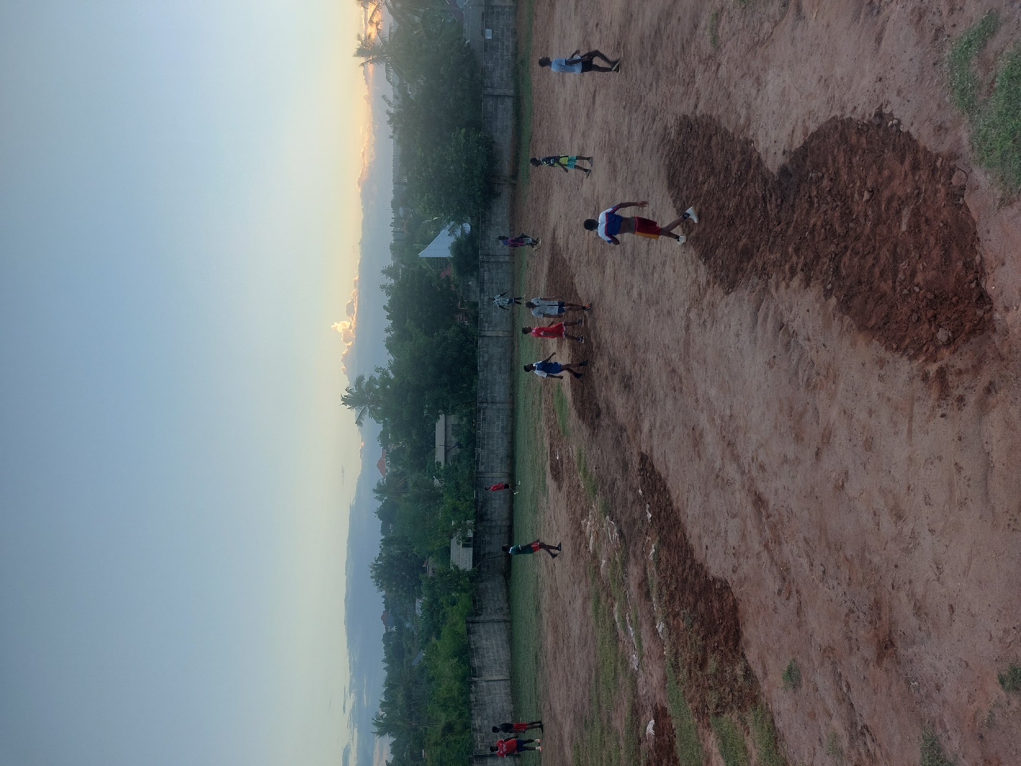 The image depicts a group of people playing on a dirt field in an area with trees and buildings, likely in a residential part of Dar es Salaam, Tanzania. The sky is light blue with a hint of yellow near the horizon. There is a wall along one side of the field. The people appear to be playing some kind of sport. The ground is mostly dirt with patches of green grass.
