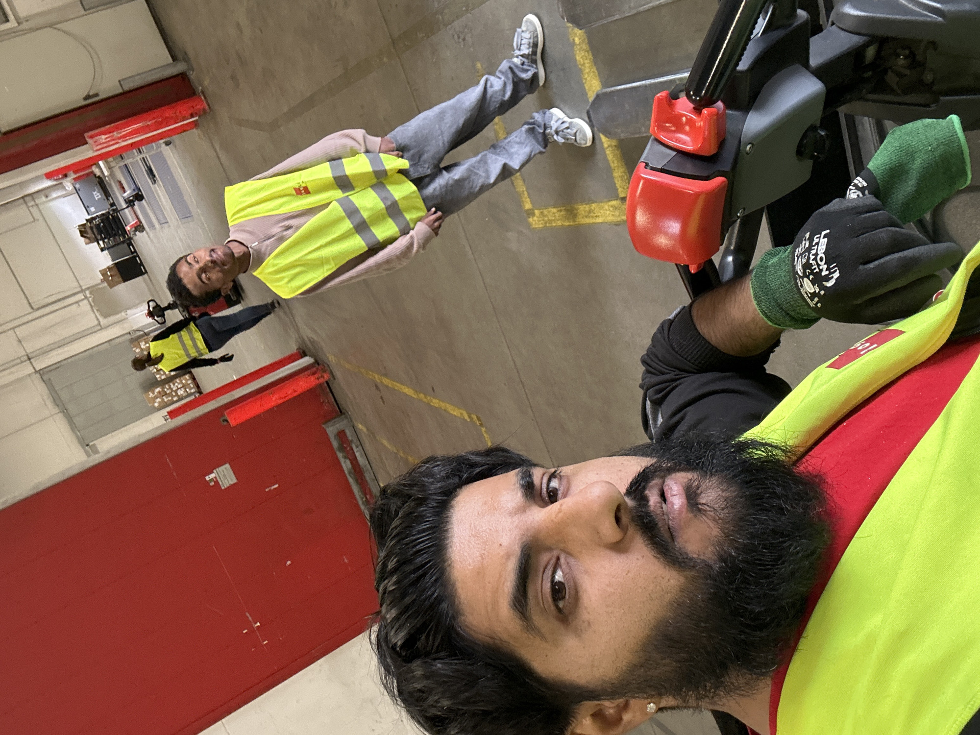The image depicts an indoor industrial or warehouse environment, possibly a logistics facility. In the foreground, a man with dark hair and a beard, wearing a red shirt and a high-visibility yellow vest, is taking a selfie. He is positioned on a piece of industrial equipment, with his gloved hand visible on the controls. In the background, another person wearing a similar high-visibility vest is walking away from the camera, and further in the distance, a third person in a yellow vest is visible near some pallets. The environment includes large red doors, concrete flooring with yellow markings, and overhead lighting. The overall impression is of a working environment within a distribution center.