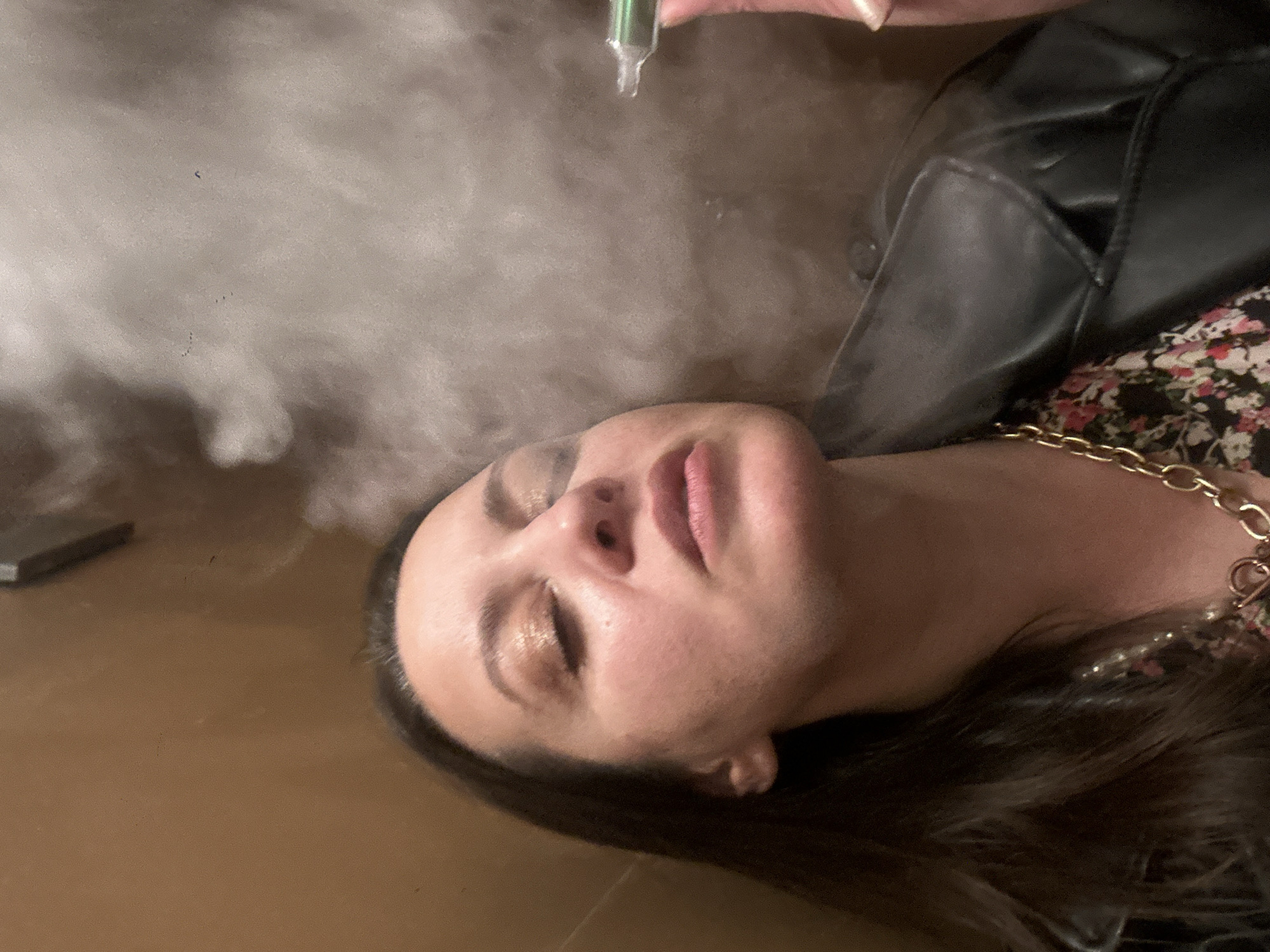 The image shows a woman with her eyes closed, lying on a surface. She has fair skin, dark hair, and is wearing makeup. She is dressed in a floral top under a black leather jacket and a gold chain necklace. A hand is holding a green vial above her face, with a vapor or smoke emanating from it. The scene is hazy with smoke.
There is no specific information in the image that indicates it was taken in Riga, Latvia.