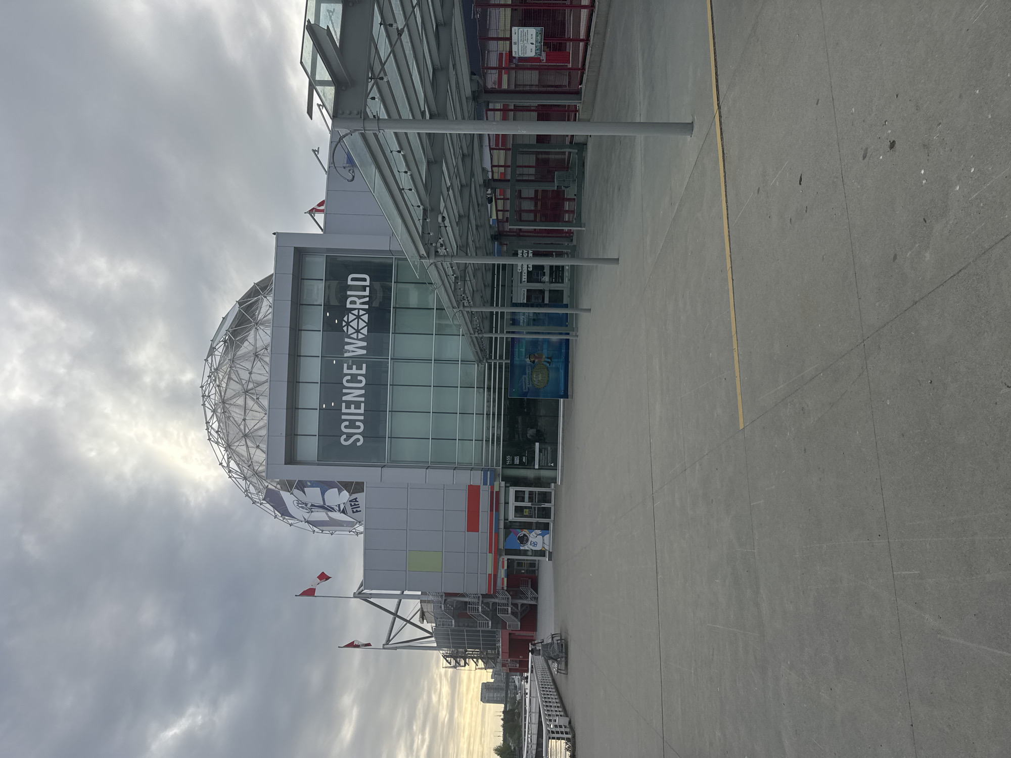 The image shows Science World, a science center in Vancouver, Canada, on a cloudy day. The iconic geodesic dome structure is visible, along with the "Science World" signage on the building. There are Canadian flags waving near the entrance. A covered walkway leads to the entrance. The foreground is a concrete plaza with yellow lines. There are no people visible in the immediate foreground, although some may be present further back near the entrance.