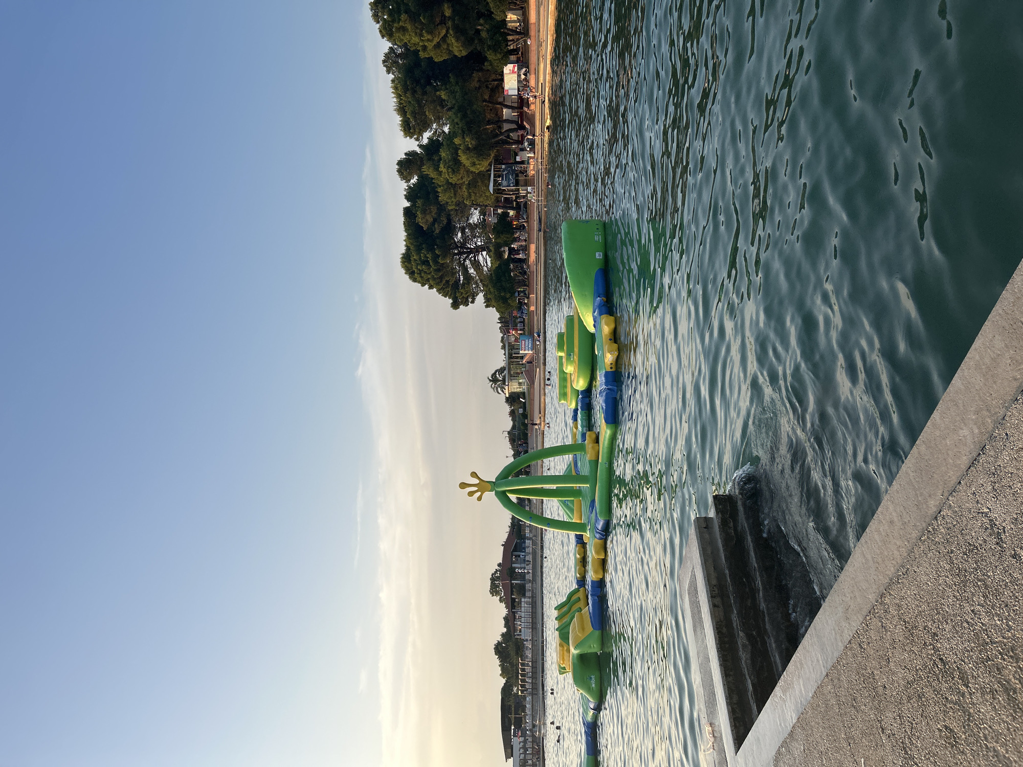 An aerial perspective captures a vibrant inflatable water park situated in the calm, turquoise waters of the Adriatic Sea, just off the coast of Katoro, Croatia. The inflatable structure, a colorful mosaic of greens and blues, features various interconnected modules, including slides, climbing structures, and platforms. A distinctive green archway topped with a yellow crown stands prominently in the center, adding a whimsical touch.
The water surrounding the park is tranquil, with gentle ripples reflecting the clear, pale blue sky. Several individuals can be seen enjoying the water park and swimming in the sea, their figures small against the expanse of the water.
In the background, a sandy beach stretches along the coastline, dotted with loungers and umbrellas, suggesting a leisurely beach day. Lush green trees, characteristic of the Mediterranean flora, form a dense backdrop to the beach. Further inland, buildings that appear to be part of a resort or leisure complex are visible, with a few scattered palm trees adding to the tropical ambiance.
The image is framed in the foreground by a concrete embankment or pier, with stone steps leading down to the water's edge. The texture of the concrete suggests it is part of a man-made structure, possibly a promenade or a dock. The overall scene conveys a sense of a serene summer day at a popular seaside destination.