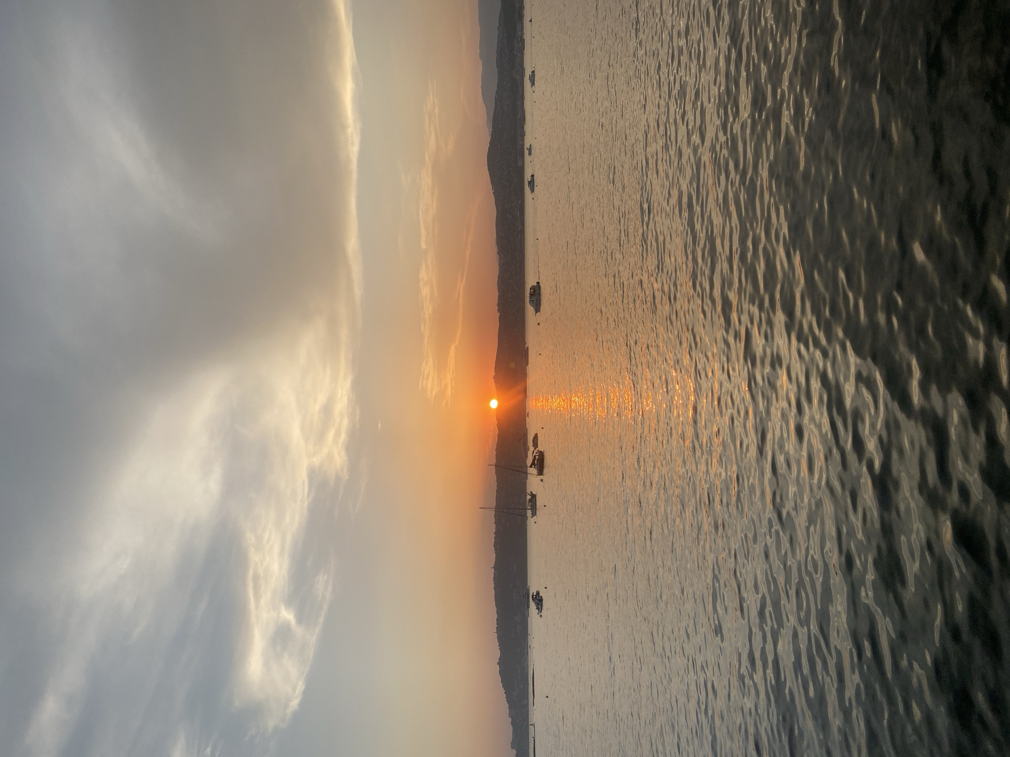 The image depicts a serene seascape at sunset, likely from the coast of Antibes, France. The sun, a bright orange orb, is low on the horizon, casting a shimmering golden path across the gently rippling water. A silhouetted coastline, dotted with trees and buildings, forms a dark band against the warm hues of the sky. Several sailboats and smaller motorboats are visible, anchored or cruising on the water, their masts and hulls faintly outlined. The sky is a gradient of soft blues, oranges, and grays, with wispy clouds catching the fading sunlight. The overall impression is one of tranquility and natural beauty, characteristic of a Mediterranean sunset.