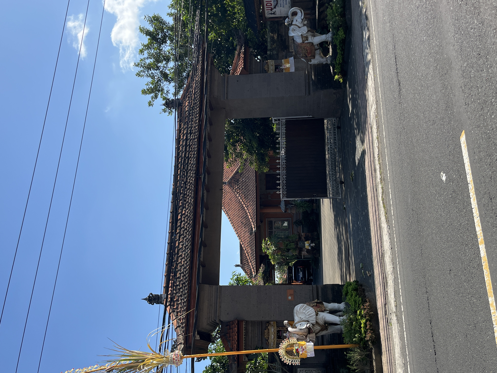 This is a photograph taken in the city of Sukawati, Indonesia, showcasing a traditional Balinese temple gate. The gate is constructed of stone and features intricate carvings, with tiled roofs. Two white elephant statues, adorned with decorative cloths, stand guard on either side of the entrance. Beyond the gate, a glimpse of the temple compound is visible, including a building with a red-tiled roof and a black metal gate. The scene is set against a clear blue sky with a few scattered clouds. The foreground of the image shows a paved road with a yellow line marking. Lush green foliage surrounds the temple structures, and power lines are visible overhead. There are no people or animals actively engaged in any activity within the frame.
