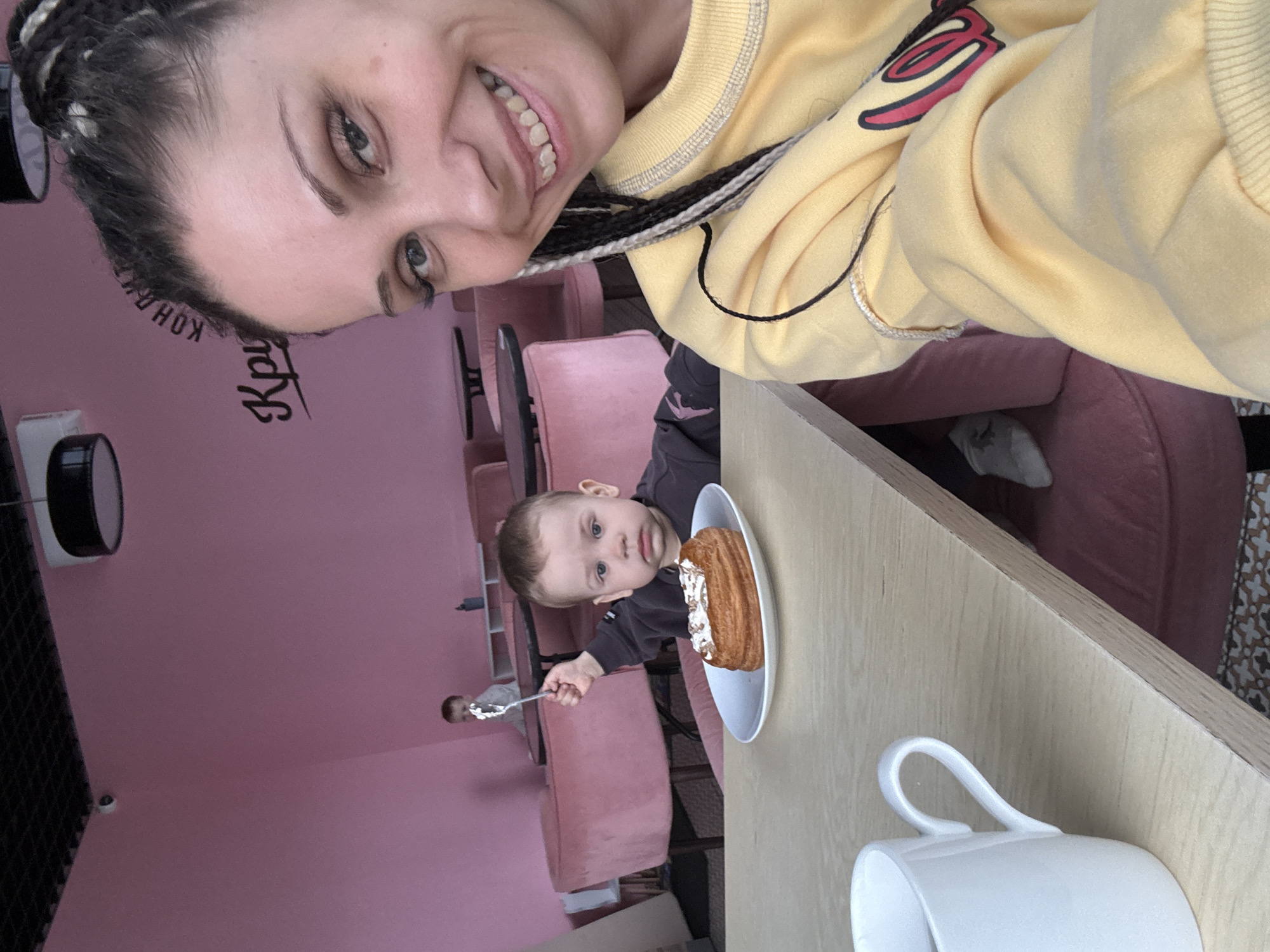 The image captures a moment in what appears to be a cafe in Rusanovo, Russia. A woman with braided hair and wearing a yellow sweatshirt is taking a selfie. A young child, possibly her son, is seated at the table with a plate containing a pastry, possibly a croissant or éclair with a white topping. The child is holding a fork, seemingly about to eat the pastry. A white mug is also visible on the table. The background features pink walls and matching upholstered chairs, typical of a cafe setting. A man is seen in the background.