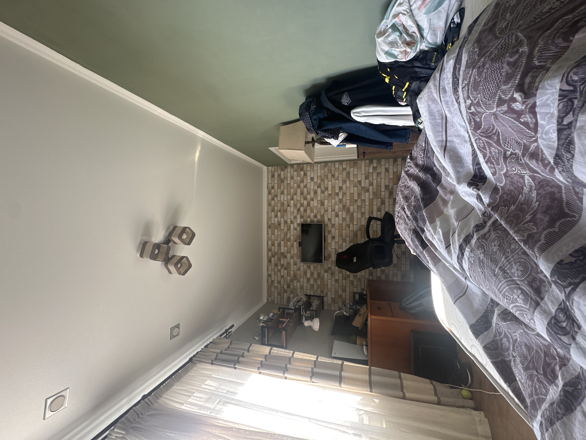 The image shows a bedroom with a bed taking up the right side of the frame, covered in a patterned duvet. To the left, there is a wall painted gray, with a unique light fixture and electrical outlets. A window with curtains is at the bottom of the frame. The back wall is brick-patterned, with a TV mounted on it. A desk and chair are visible near the back wall. Piles of clothes and other items are present on top of a dresser, also near the back wall. The ceiling is two-toned, with gray and green sections. The location of the scene is unknown.